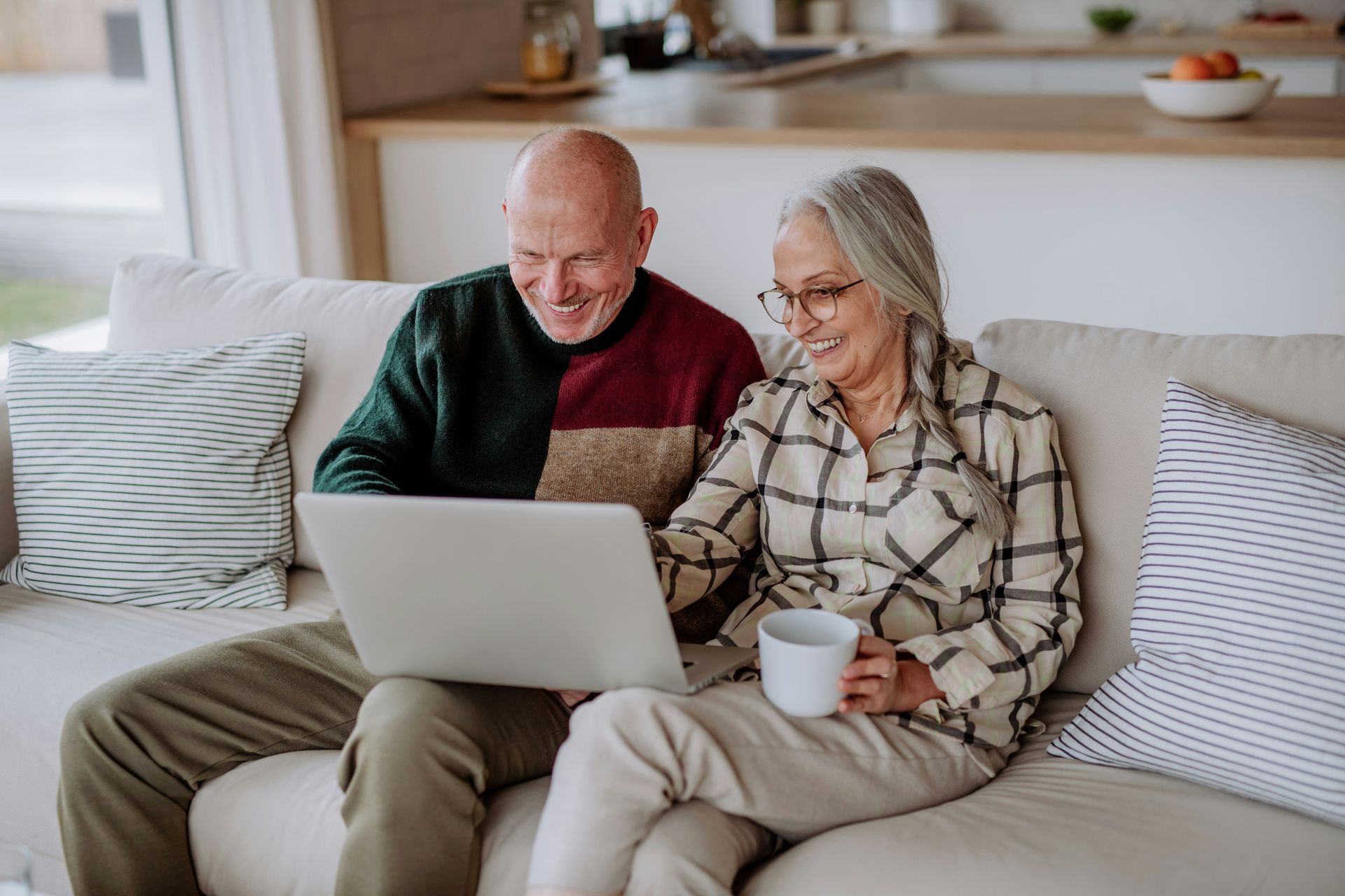 Older couple looking at a laptop together on a couch; one holding a mug.