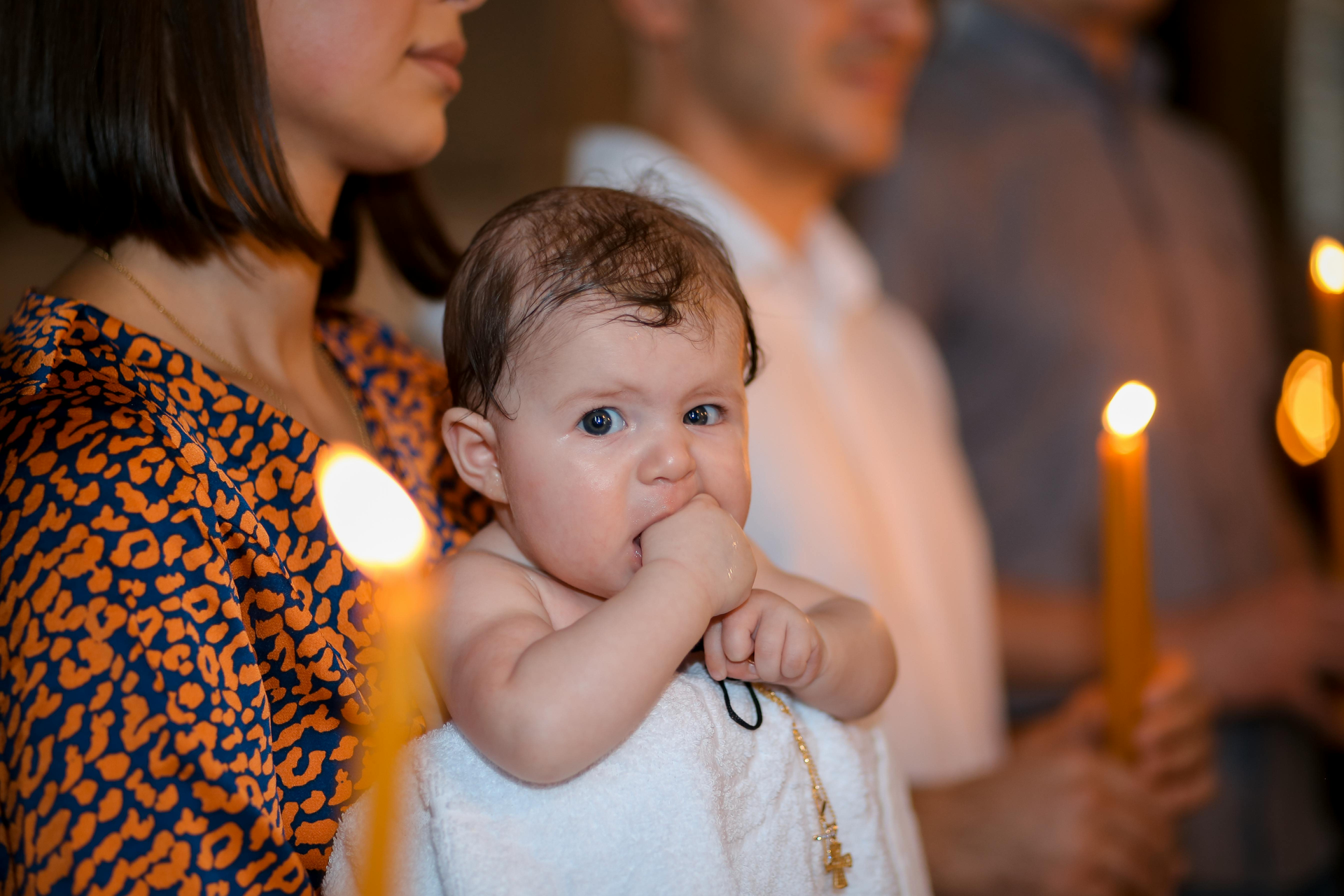 Baby with wet hair, sucking fingers, held during a baptism ceremony with candles.