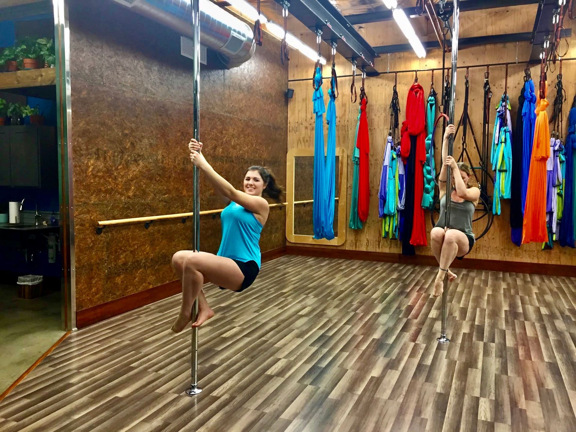 Two women pole dancing in a studio with colorful hanging silks and wood floors