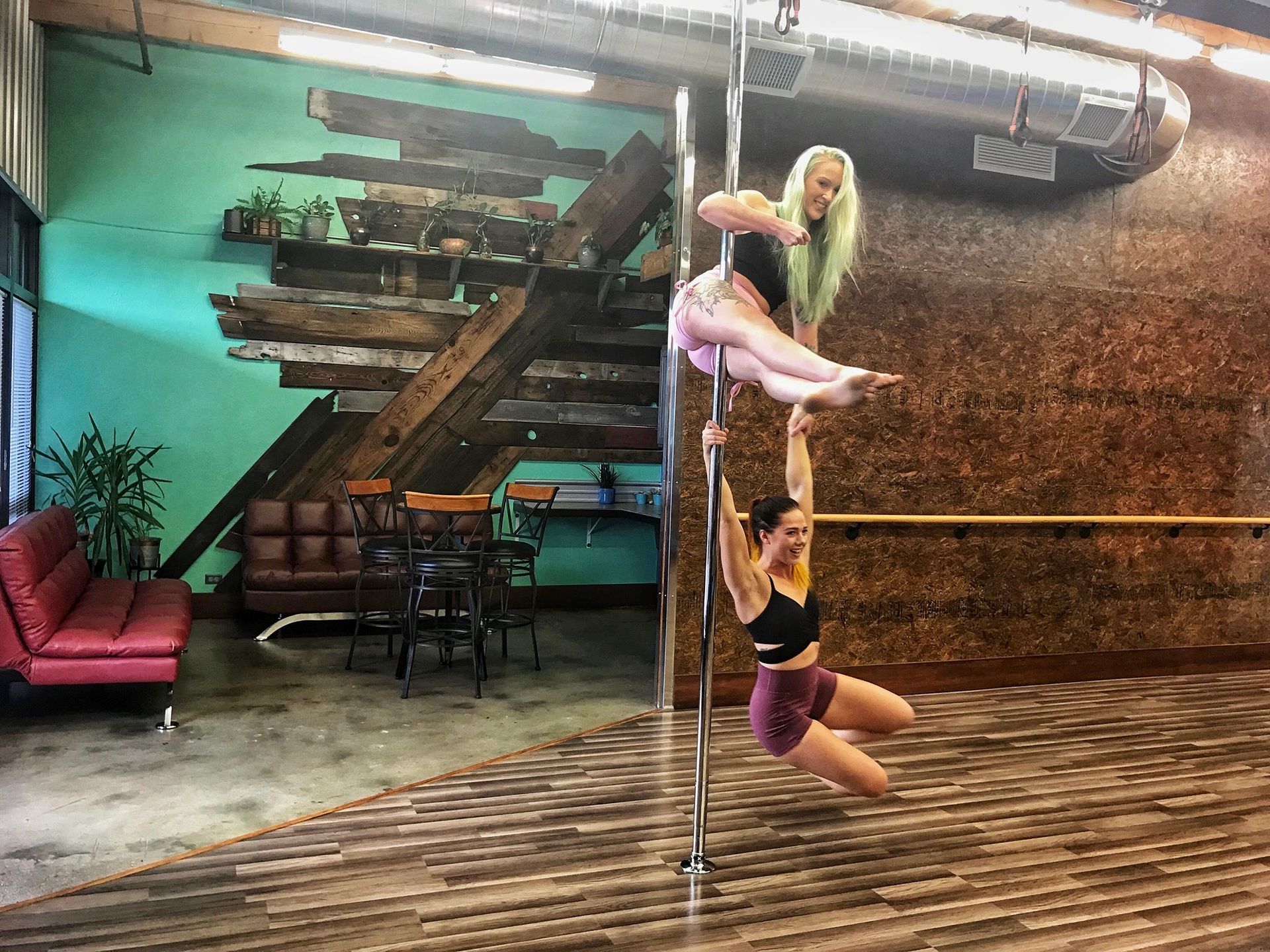 Two people practicing pole dance in a studio with teal walls and industrial decor