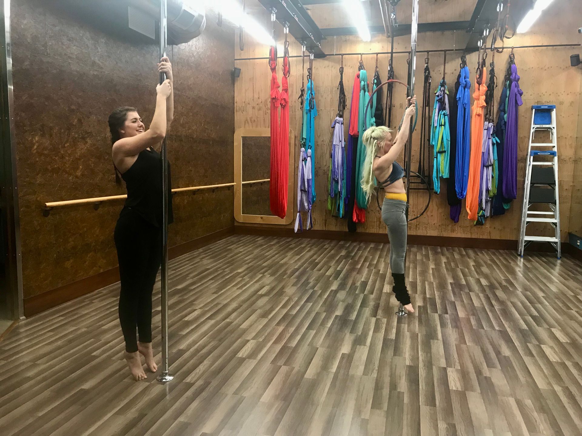 Two women pole dancing in a studio with hanging silks and a ladder in the background