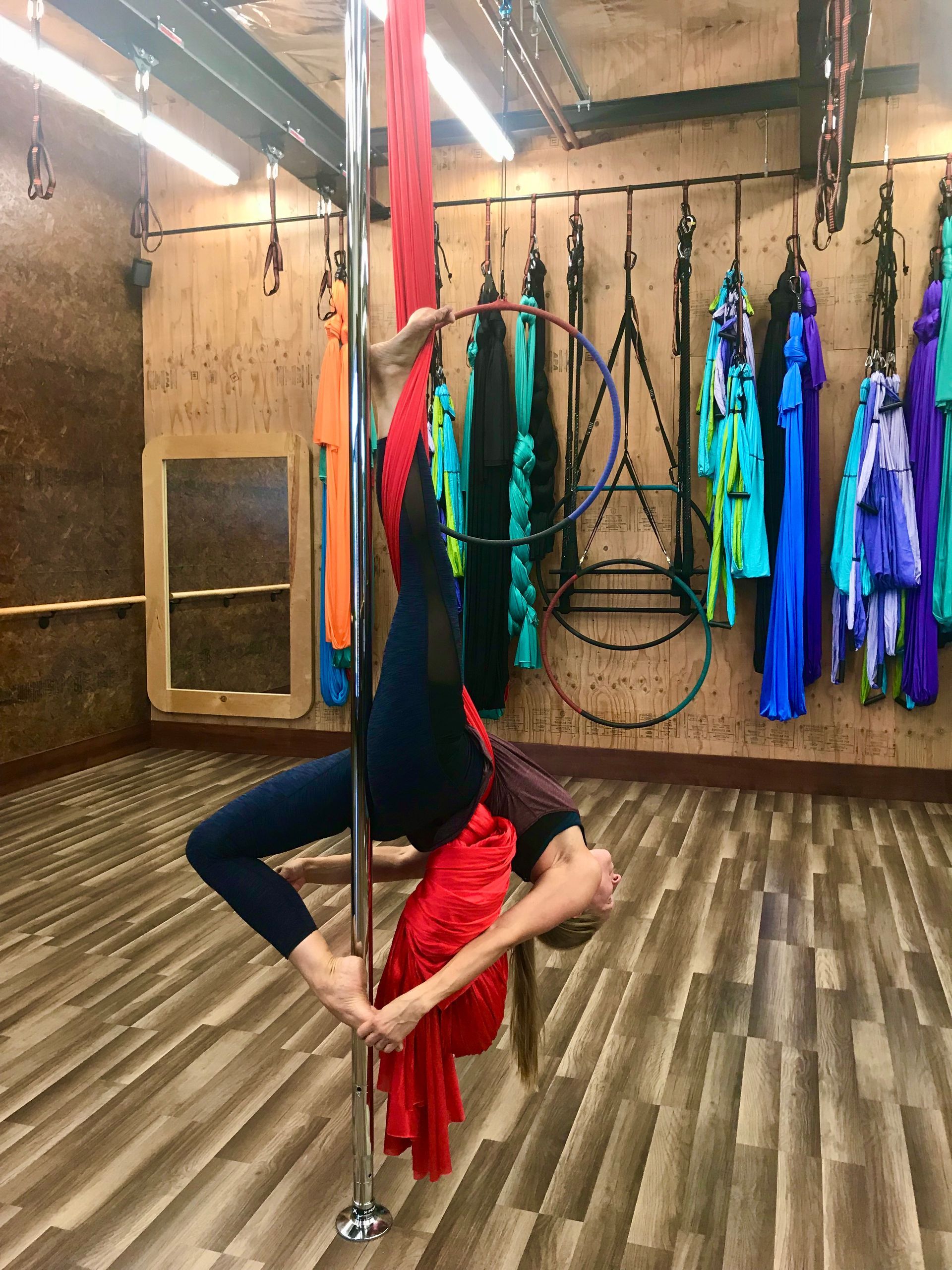 Person hanging upside down from red aerial silks in a studio, with colorful hammocks and ropes behind.