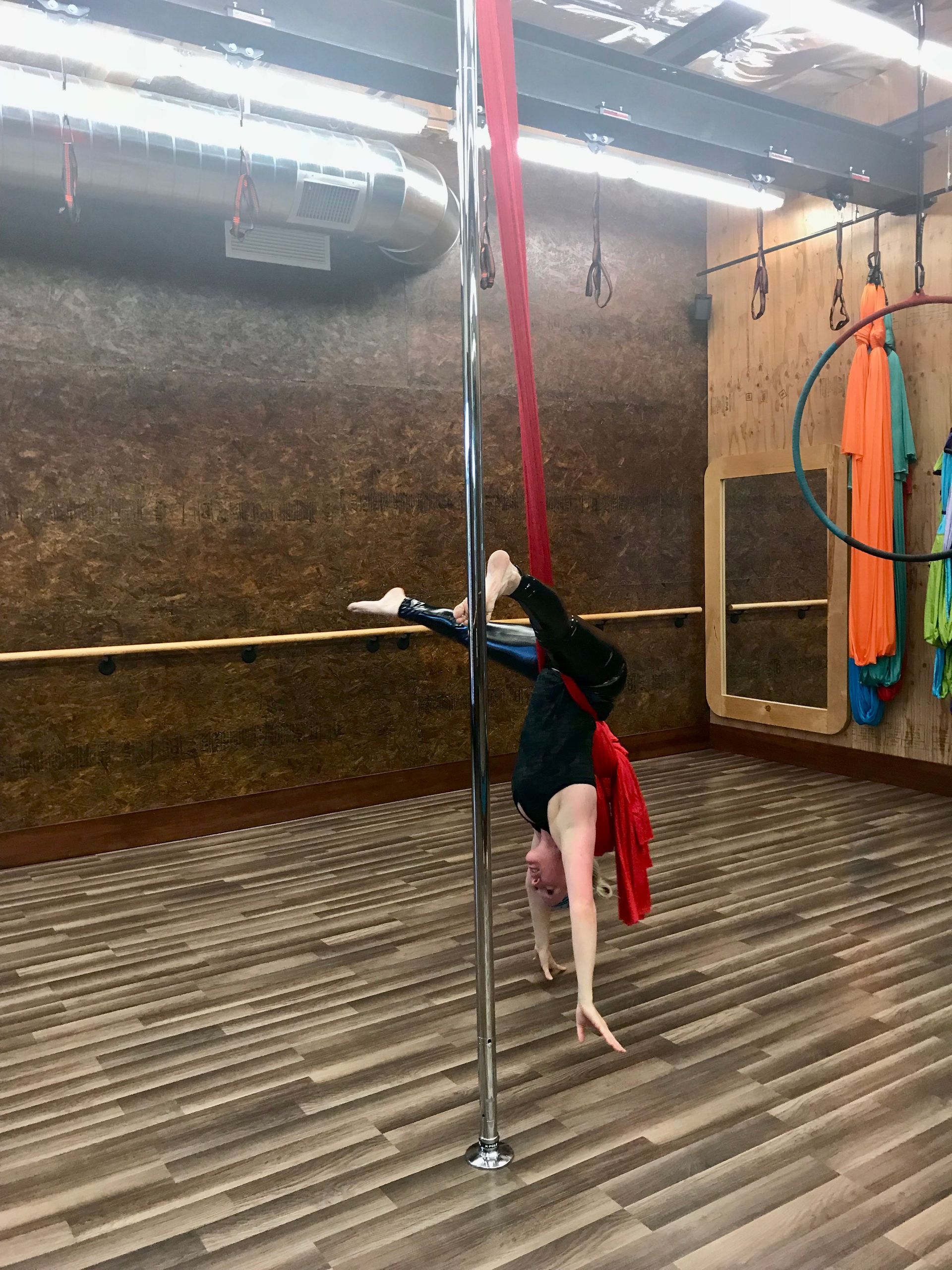 Person performing a pole dance move in a studio, wearing black and red, with hanging silks nearby.