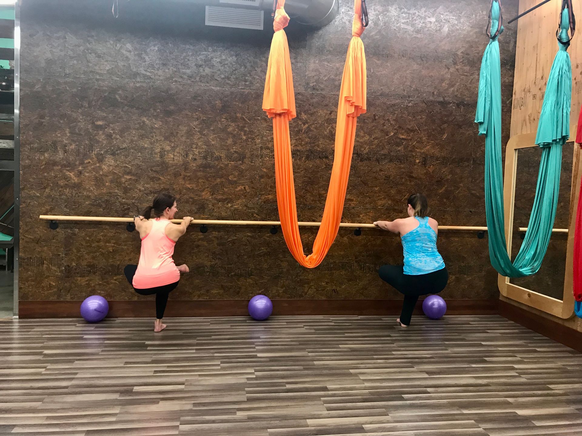Two people stretching at a barre in a studio with hanging silk hammocks and exercise balls.