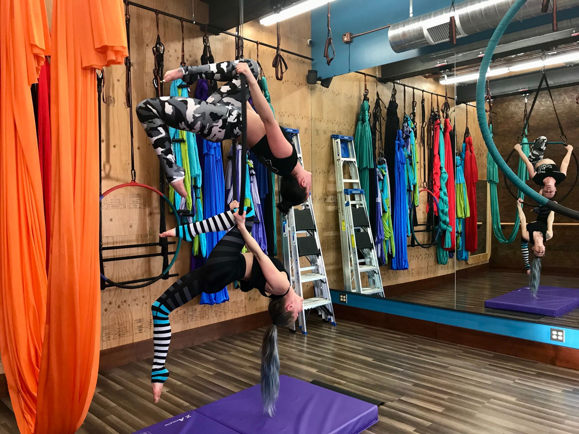 Aerial yoga studio with a woman inverted on a purple mat, mirrors, hammocks, and hanging silks around her.