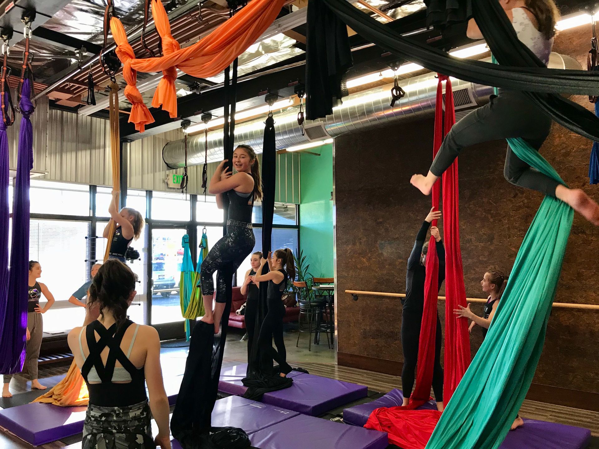 People doing aerial silk exercises in a bright studio with purple mats and colorful hanging fabrics