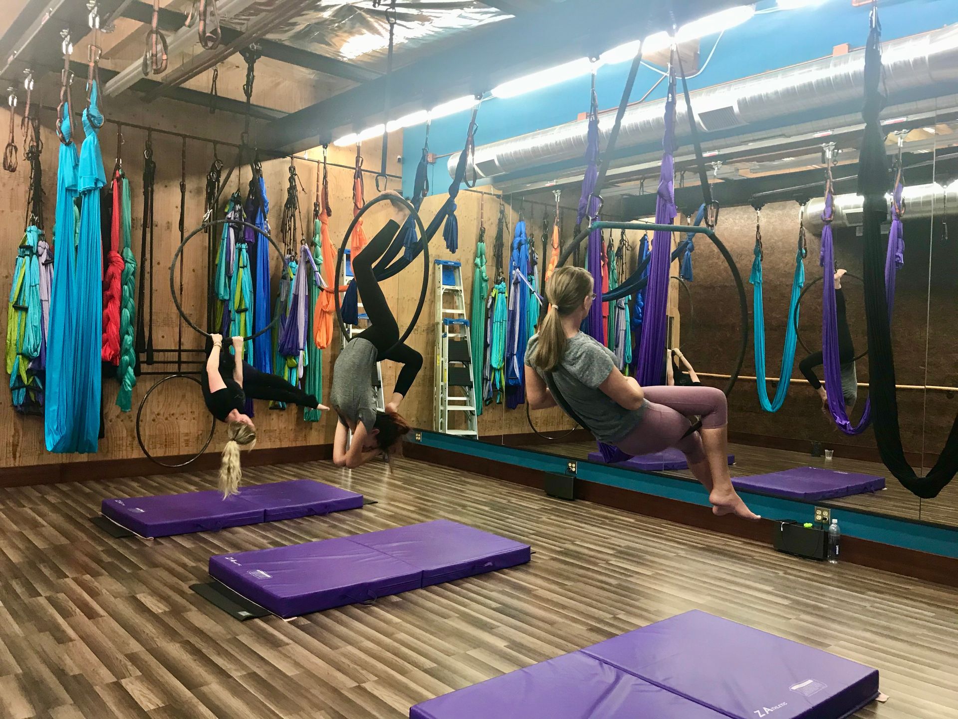 Three people using aerial yoga hammocks in a studio with purple mats and wood floors
