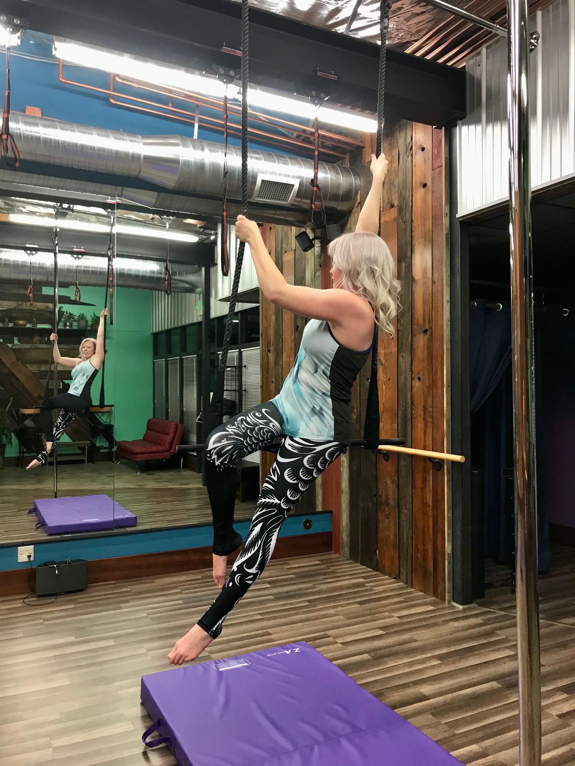 Woman practicing aerial silks in a studio, hanging from fabric above purple mats and a mirrored wall.