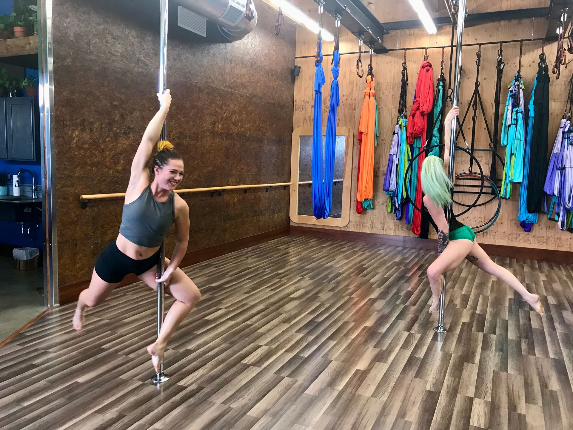 Two women in aerial silks poses in a studio with hanging colorful fabric and wooden floors