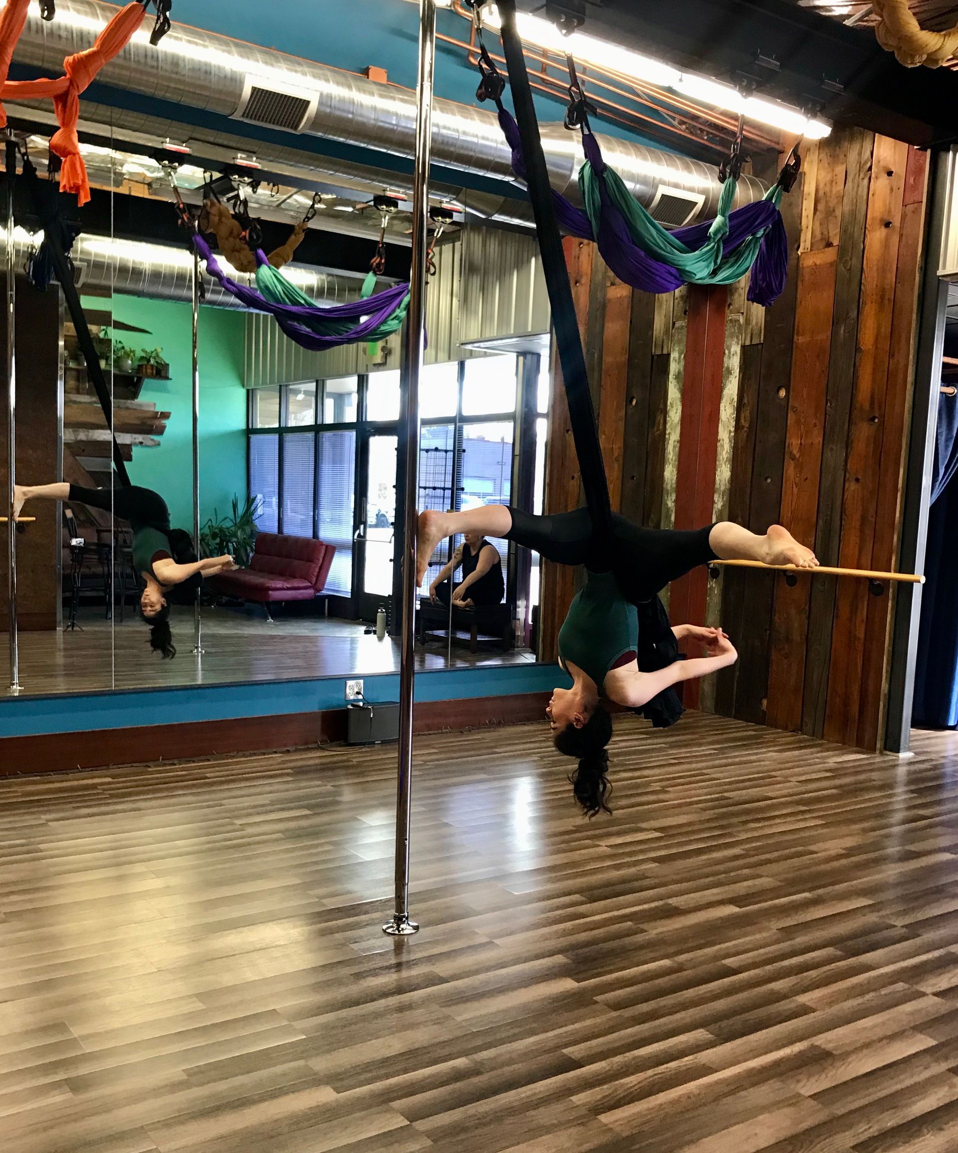 Person performing an inverted pole dance move in a studio with mirrors and wooden floors