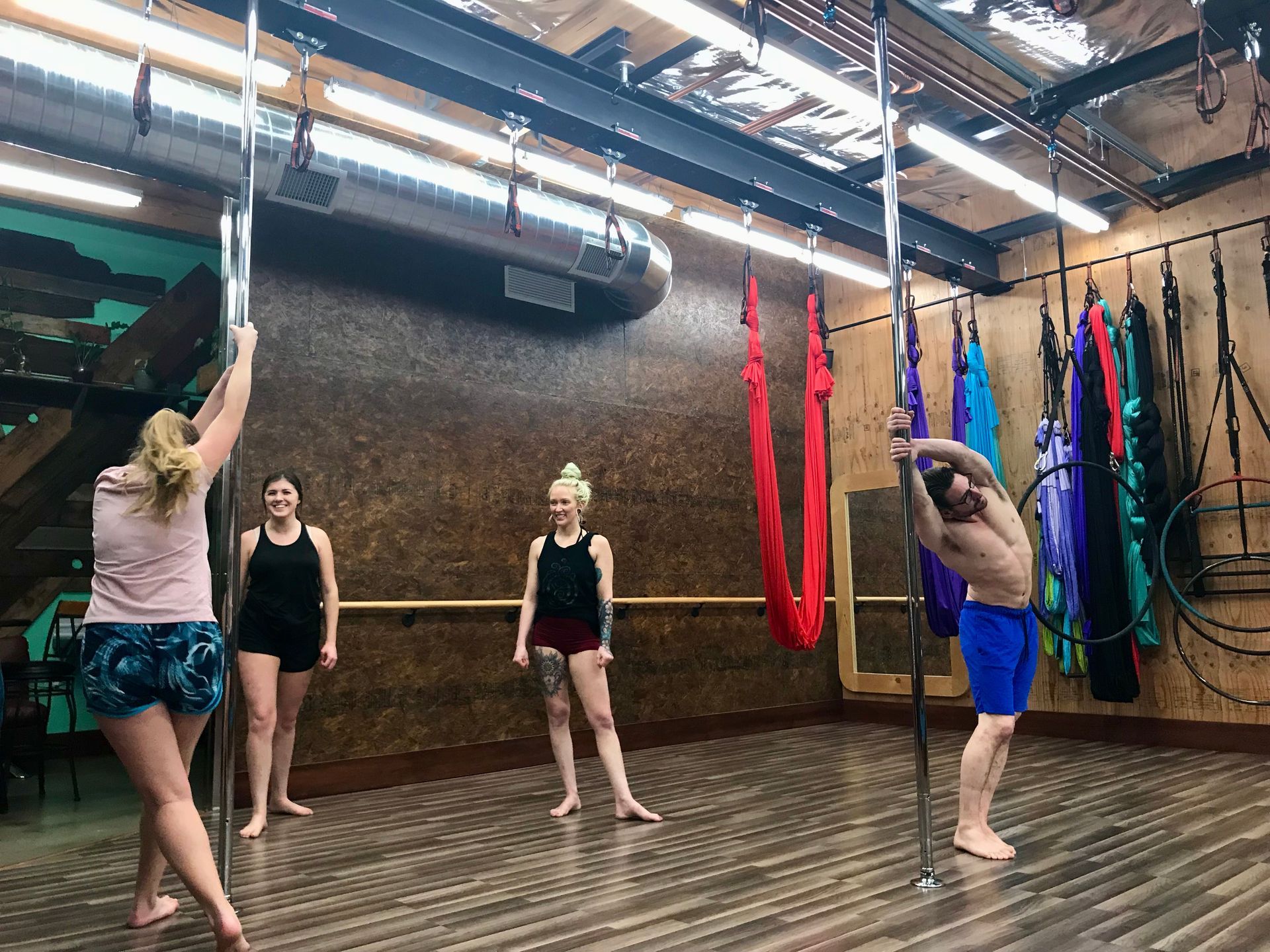 People stretching and hanging from aerial silks in a studio with wood floor and exposed ceiling beams