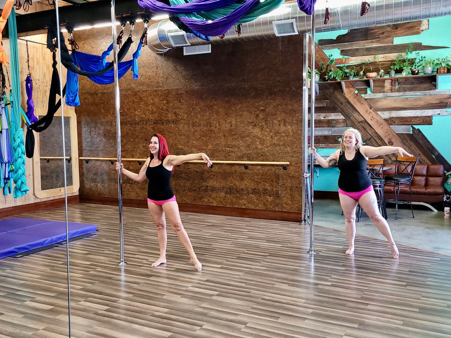 Two people in dance attire practicing in a mirrored studio with wooden floors and aerial silks overhead