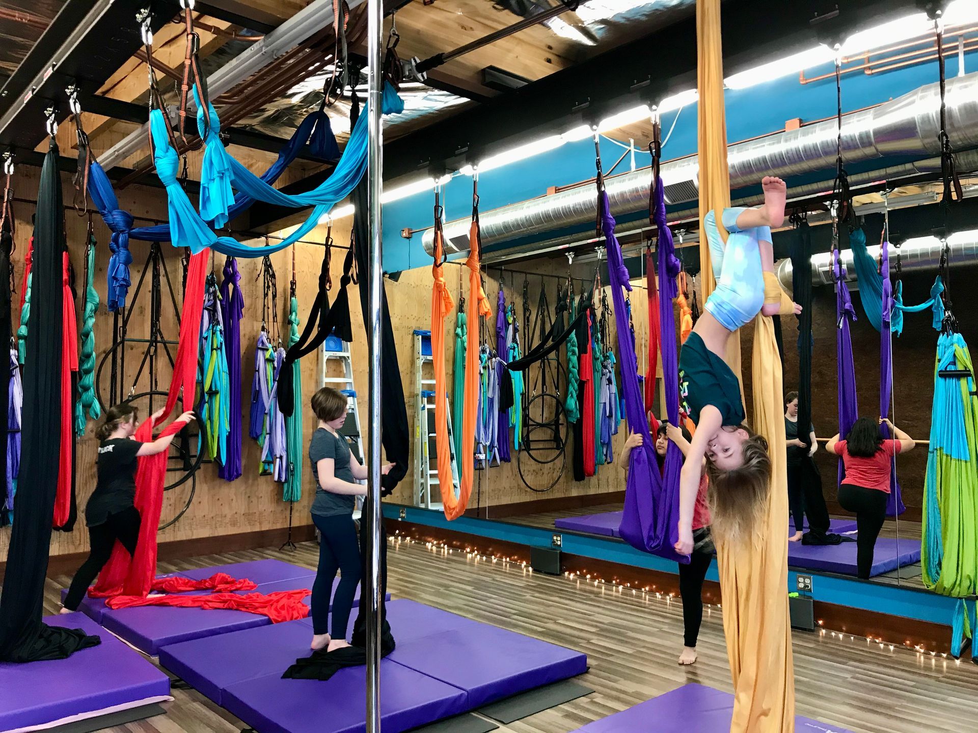 People doing aerial yoga on colorful hammocks in a studio with purple mats and mirrors