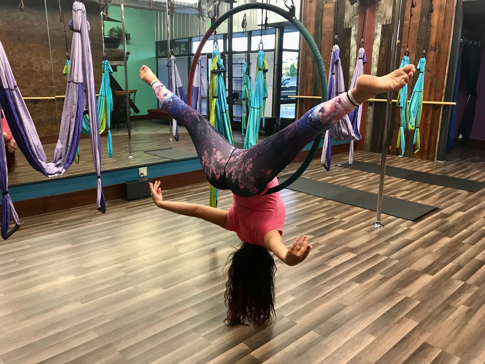 Inverted aerial performer in a studio, holding a wide split on a mat with hanging silks and poles behind.
