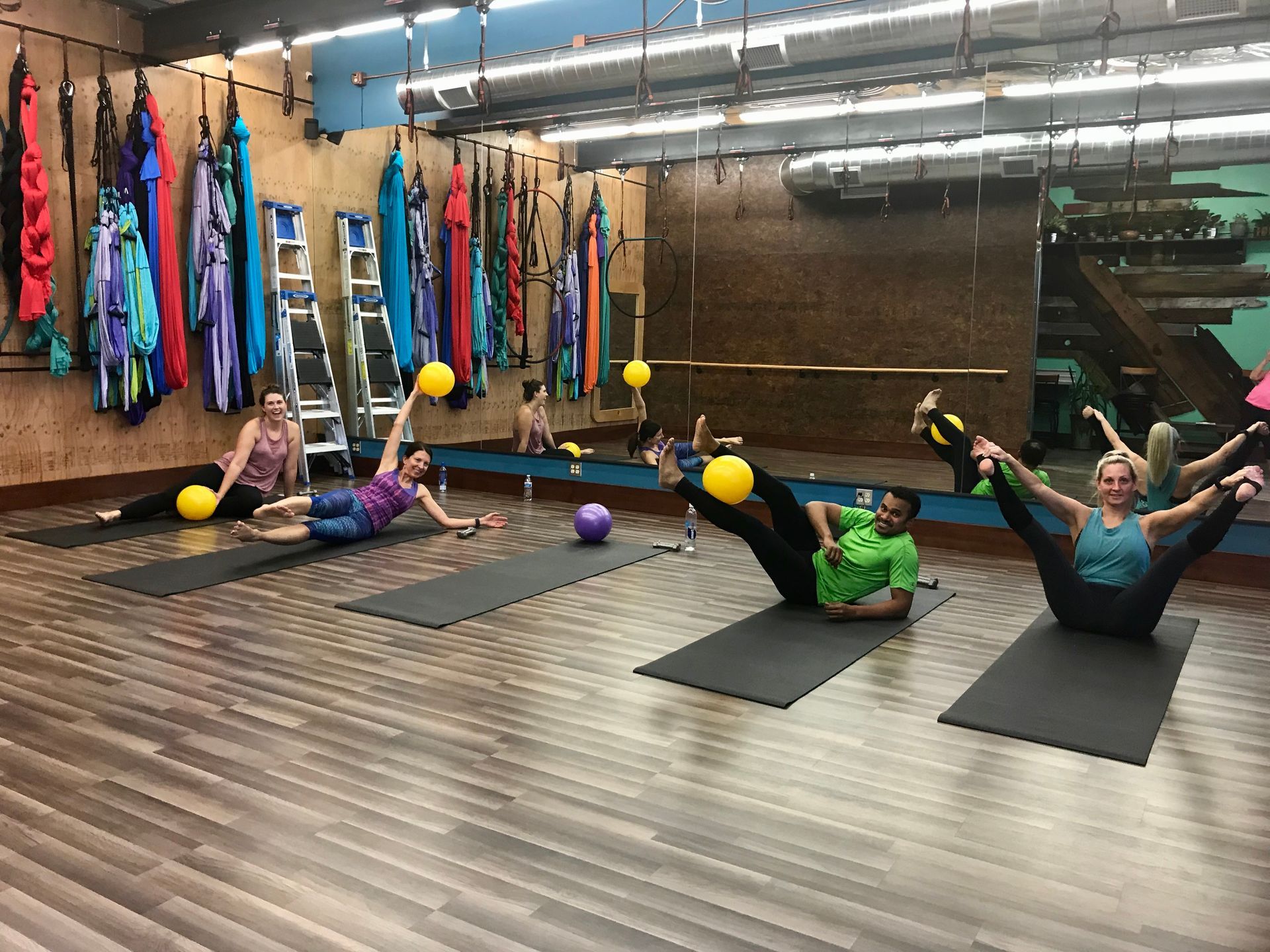 People exercising on mats in a yoga studio with colorful hanging fabrics and exercise balls