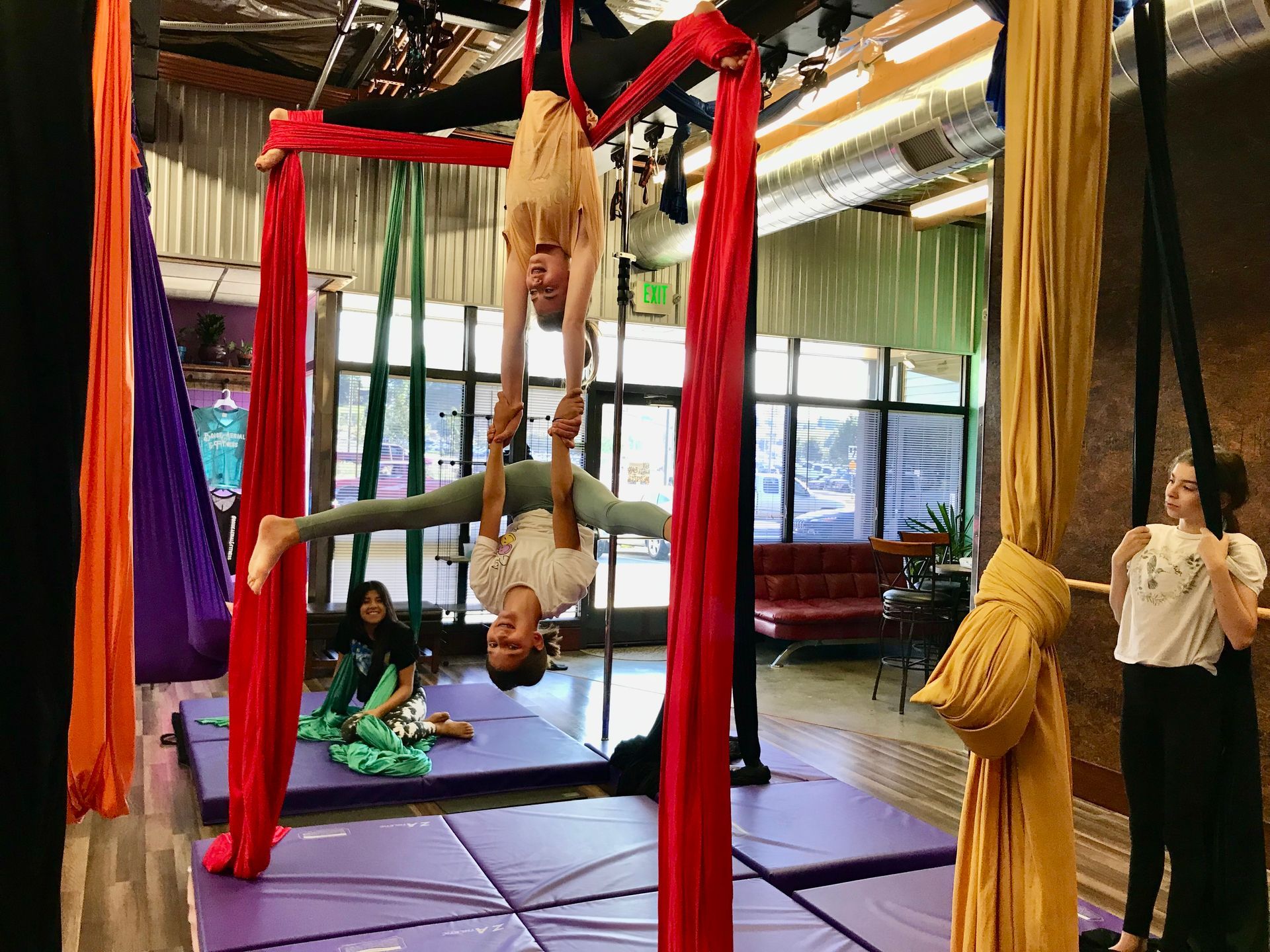 People practicing aerial yoga on red silks in a studio with purple mats and large windows