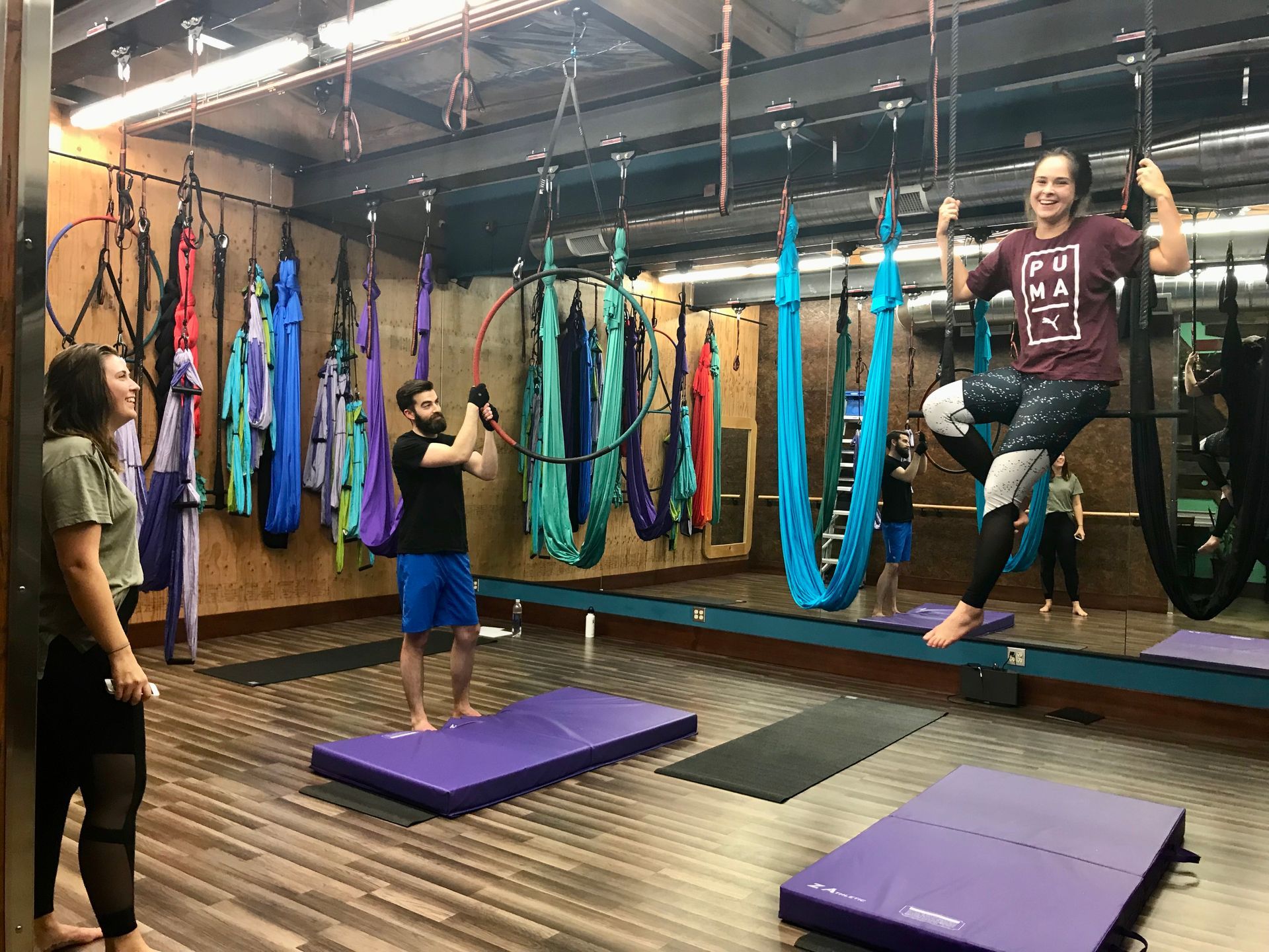 Indoor aerial yoga class with people on colorful hammocks and purple mats in a studio
