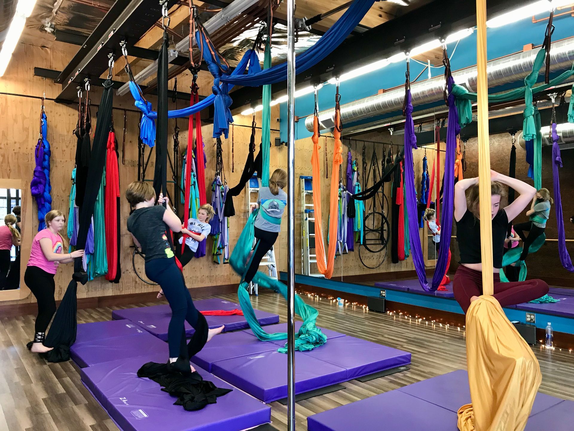People practicing aerial yoga in a studio with colorful hanging silks and purple mats.