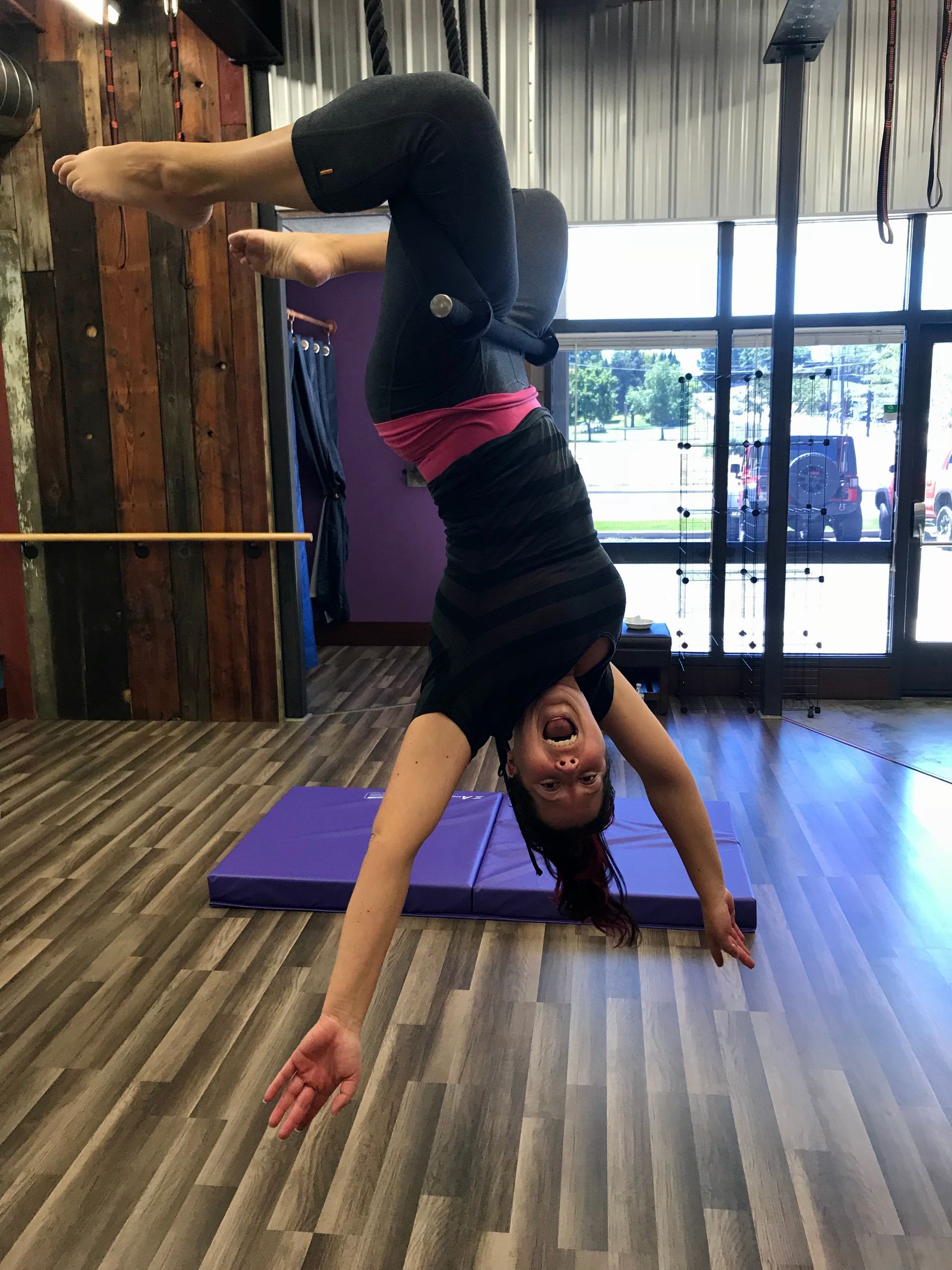 Person doing a one-handed handstand in a studio with wood floors and large windows.