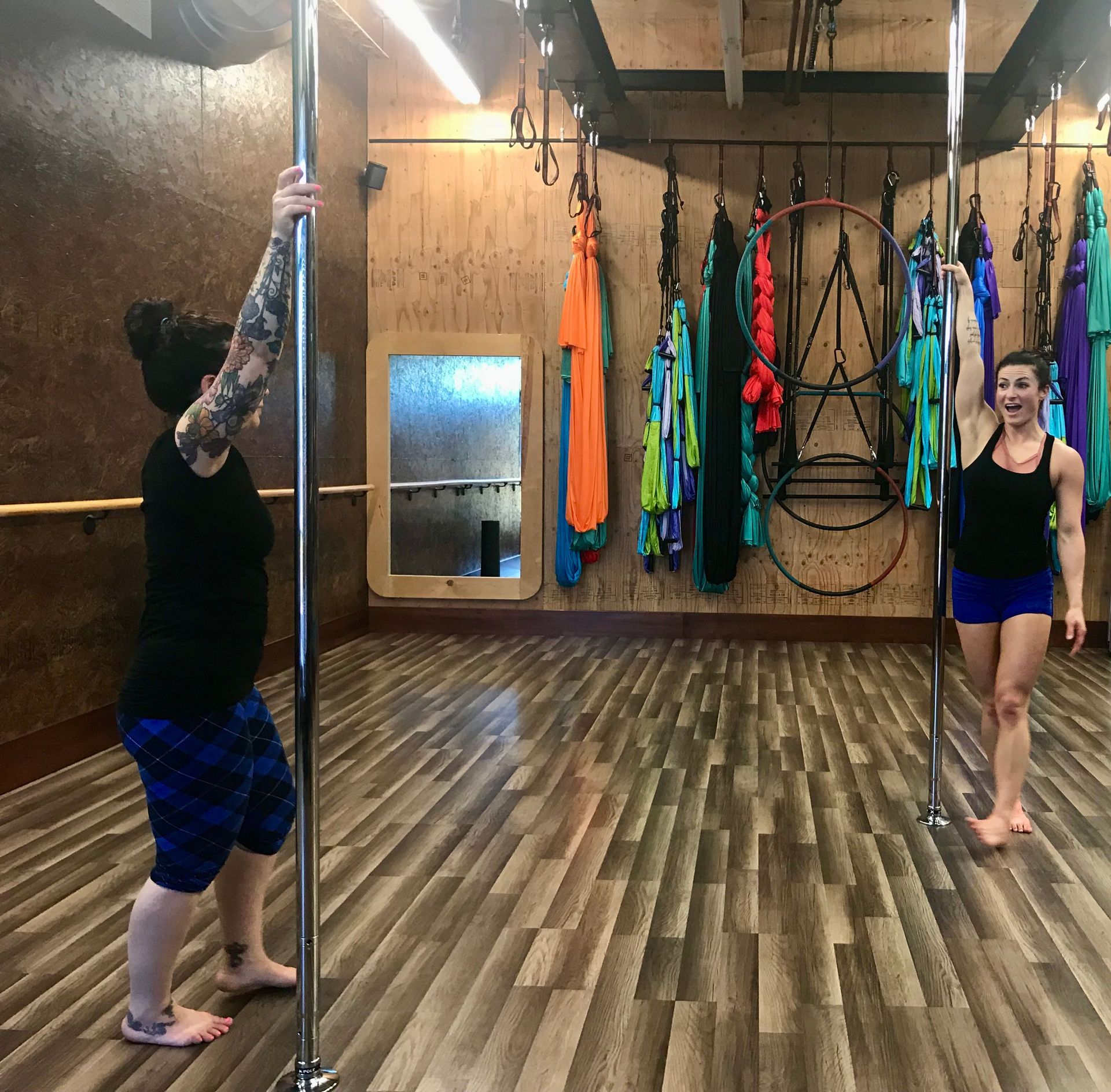 Two people in a pole dance studio, one gripping a pole and one standing nearby among aerial silks.
