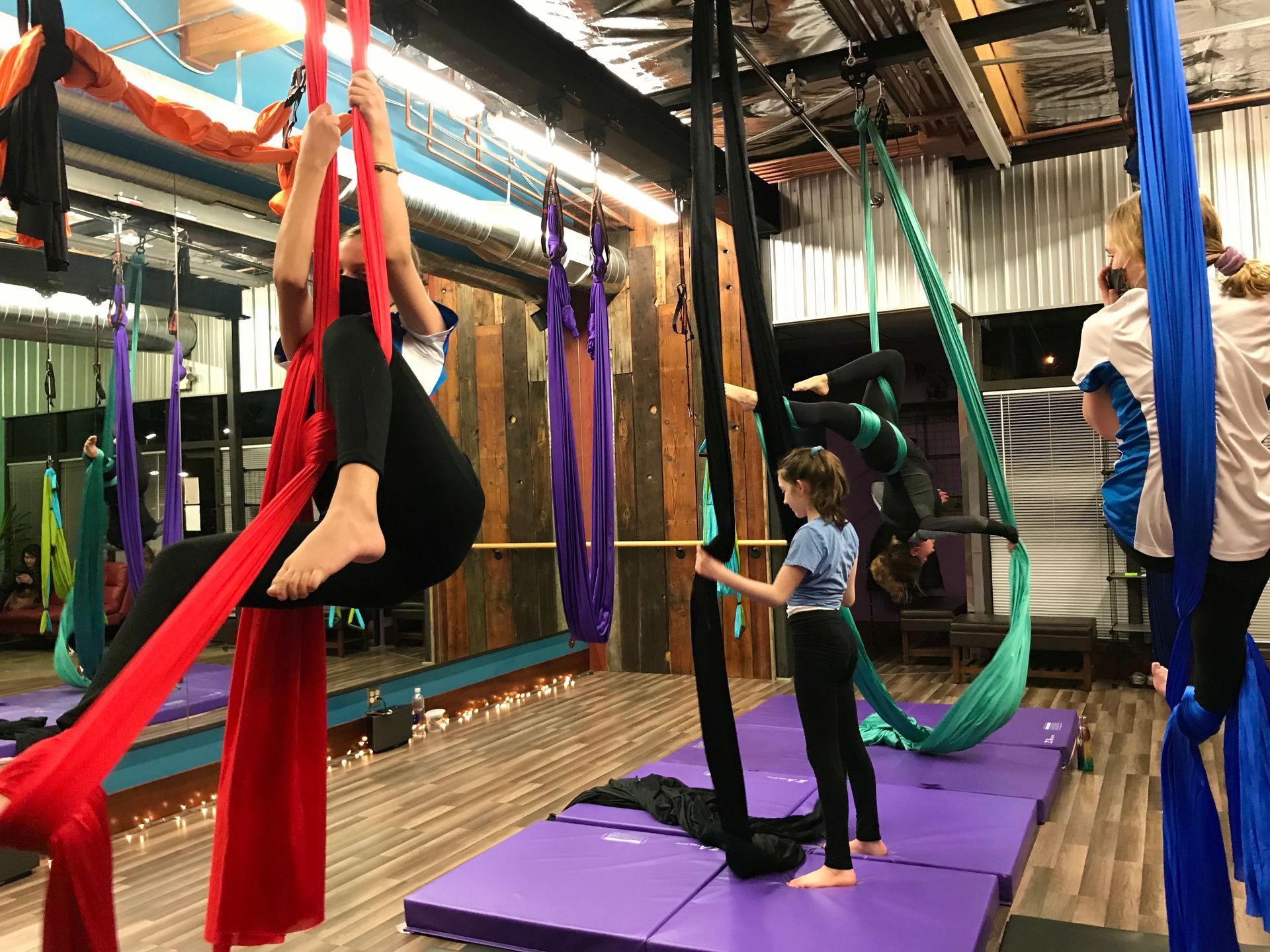 People practicing aerial yoga on colorful hammocks in a studio with purple mats.