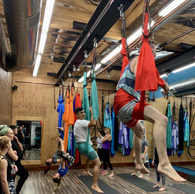 People practicing aerial yoga in colorful hammocks in a studio with wood walls and exposed ceiling beams