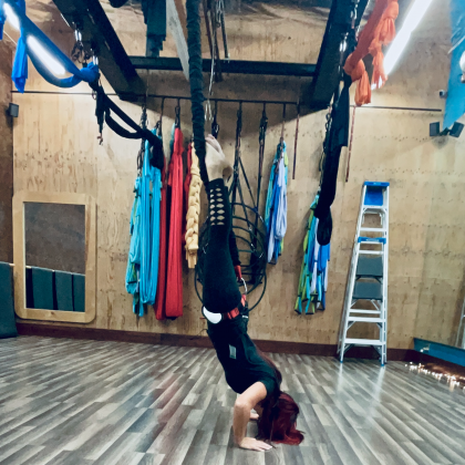 Person doing a headstand in a studio with hanging aerial silks and a ladder in the background