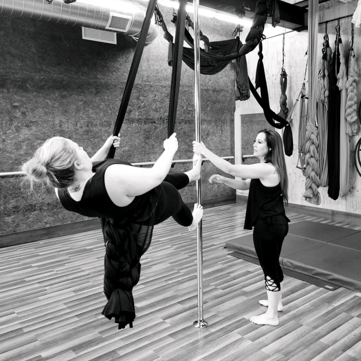 Two people practicing aerial hammock in a gym, one suspended sideways while the other steadies the fabric.