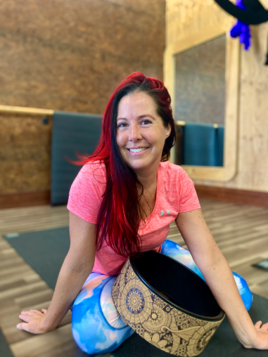 Smiling person seated on a yoga mat beside a patterned drum in a studio, wearing a pink top and blue leggings