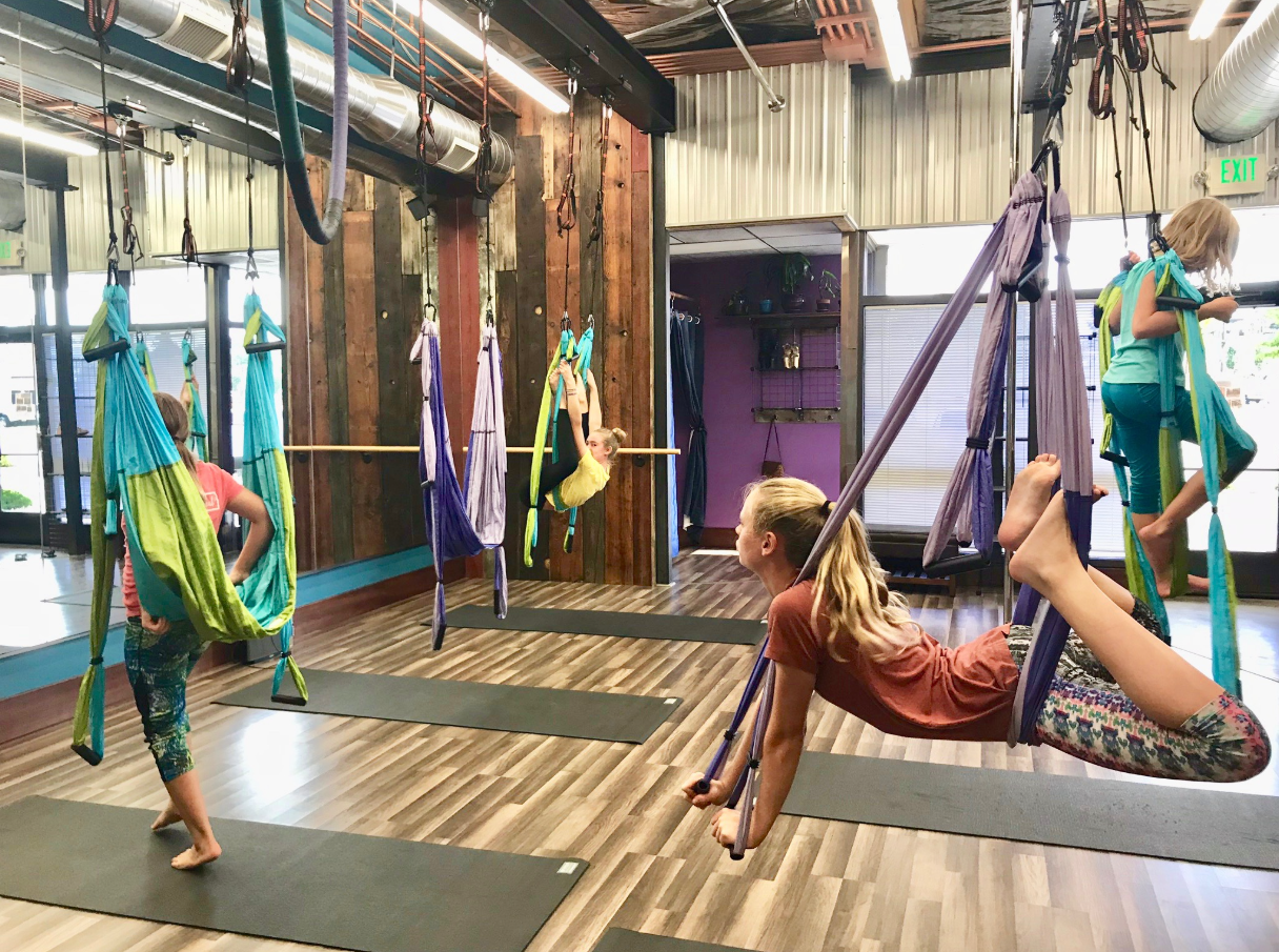 People practicing aerial yoga in a studio, suspended in colorful hammocks on mats.
