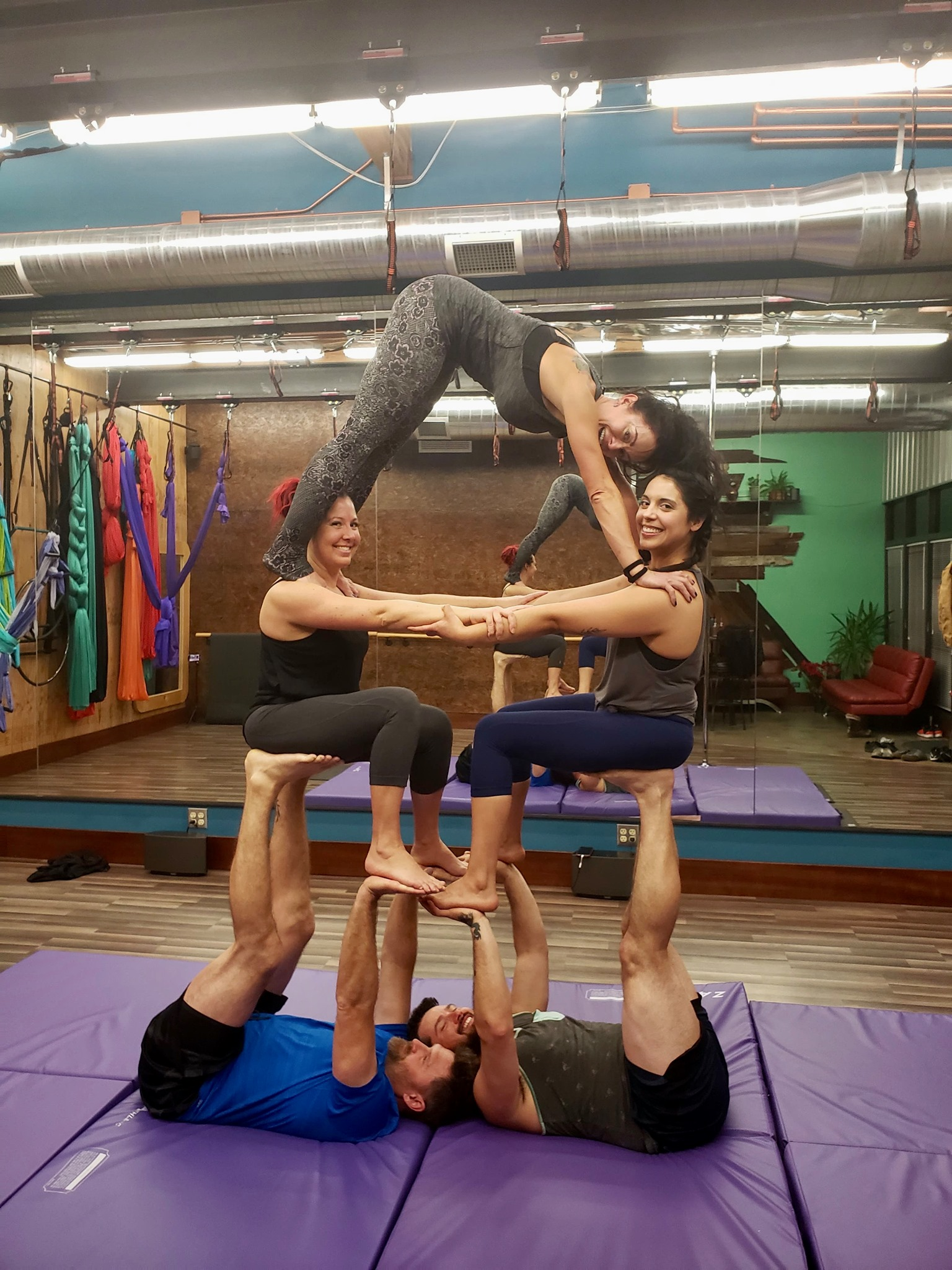 Four acrobats forming a human pyramid in a studio, balanced on mats with colorful silks hanging behind.