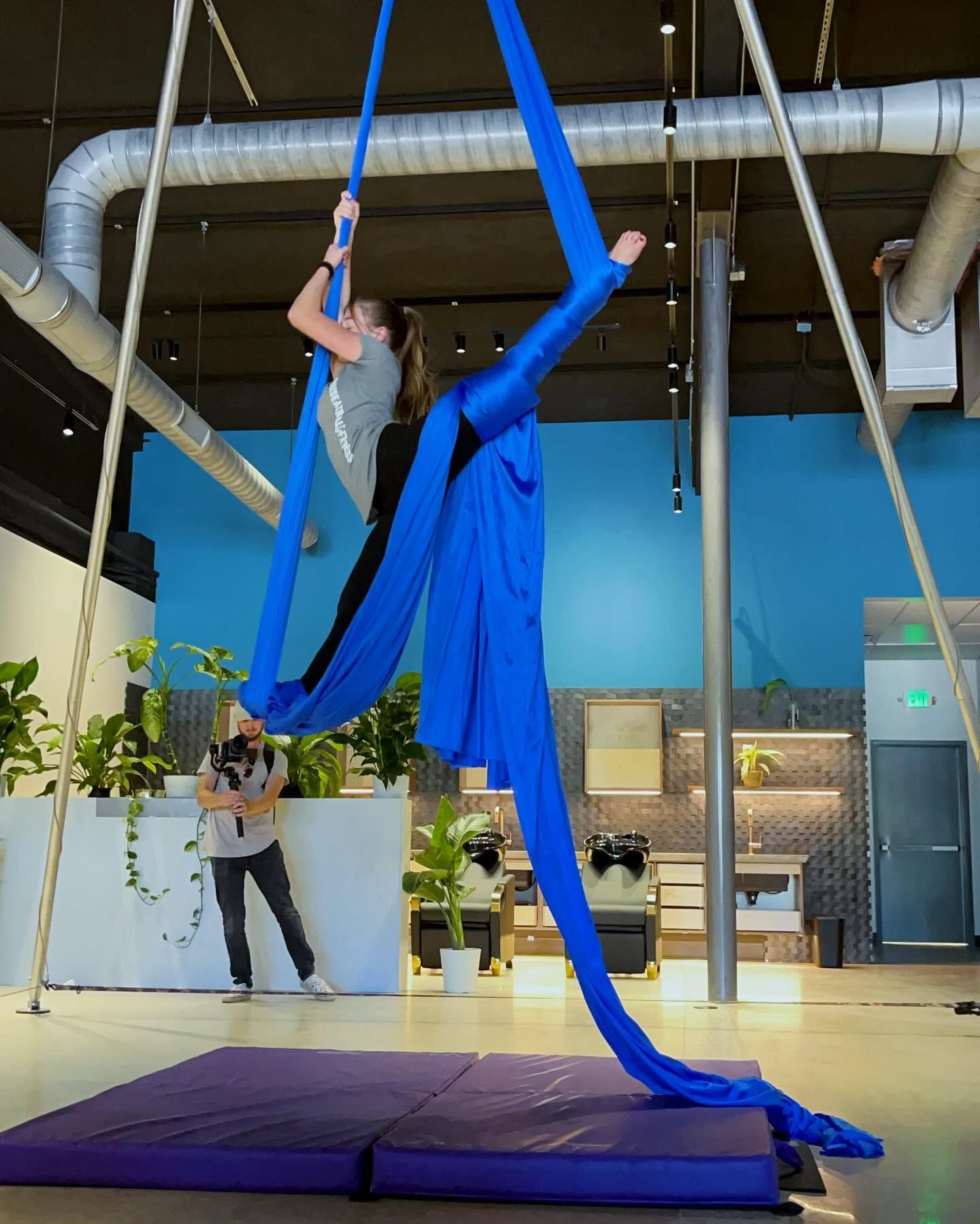 Person performing aerial silk pose in a bright indoor studio over purple mats