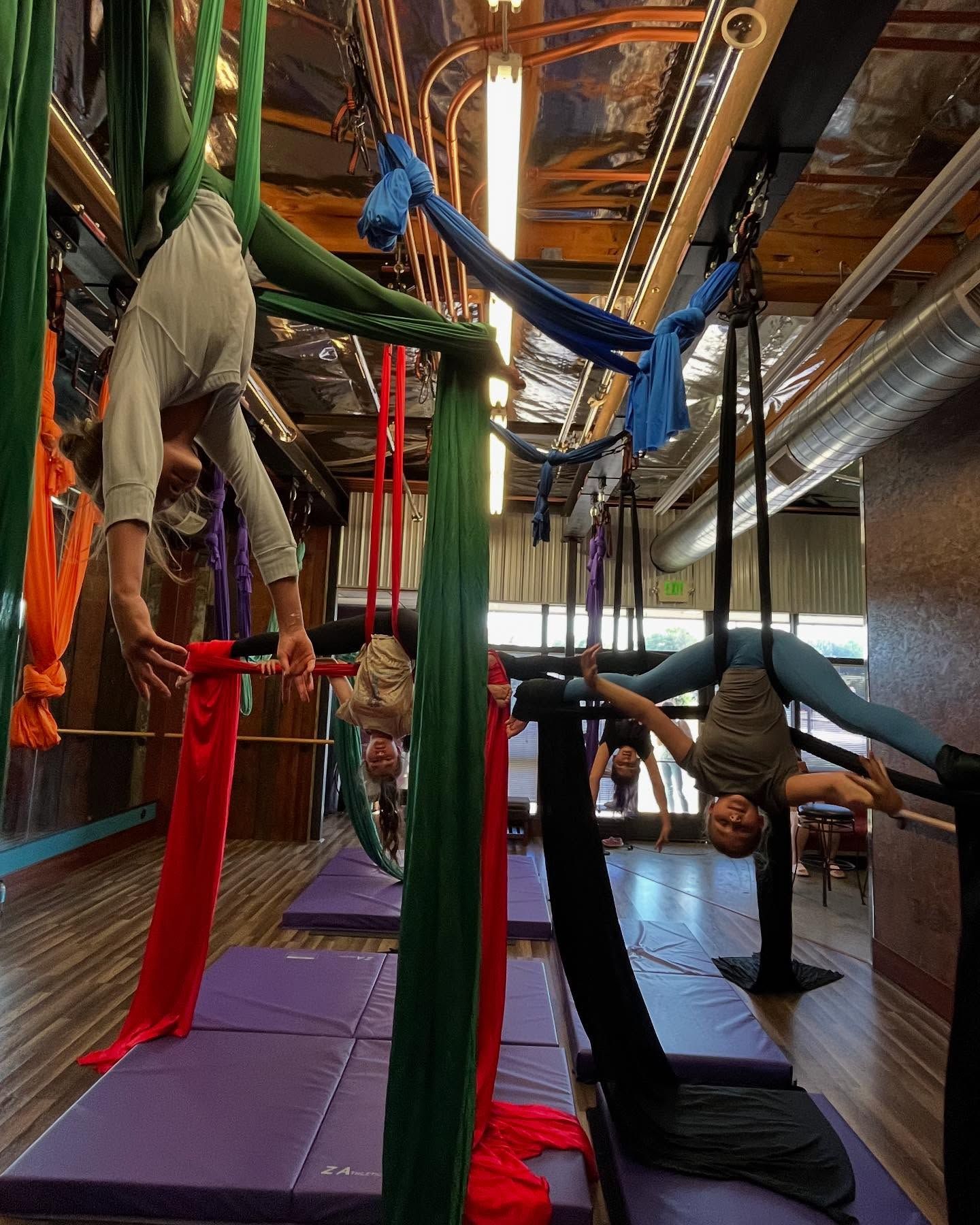 People suspended in colorful aerial yoga hammocks in a studio with mirrored walls and wood flooring
