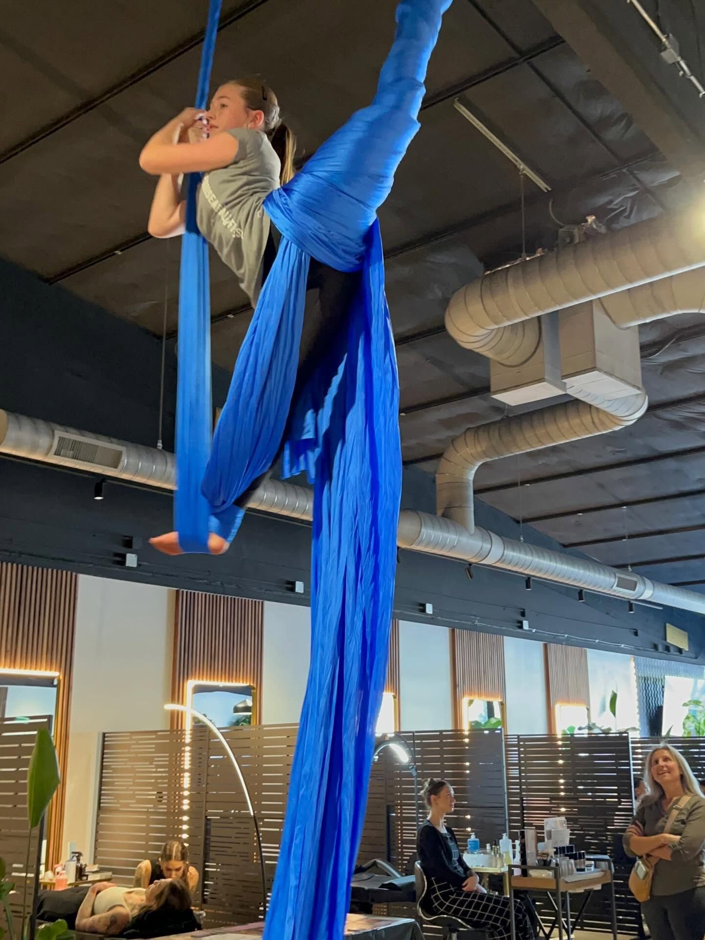 Aerial silk performer in blue fabric poses upside down in an indoor studio with onlookers below.