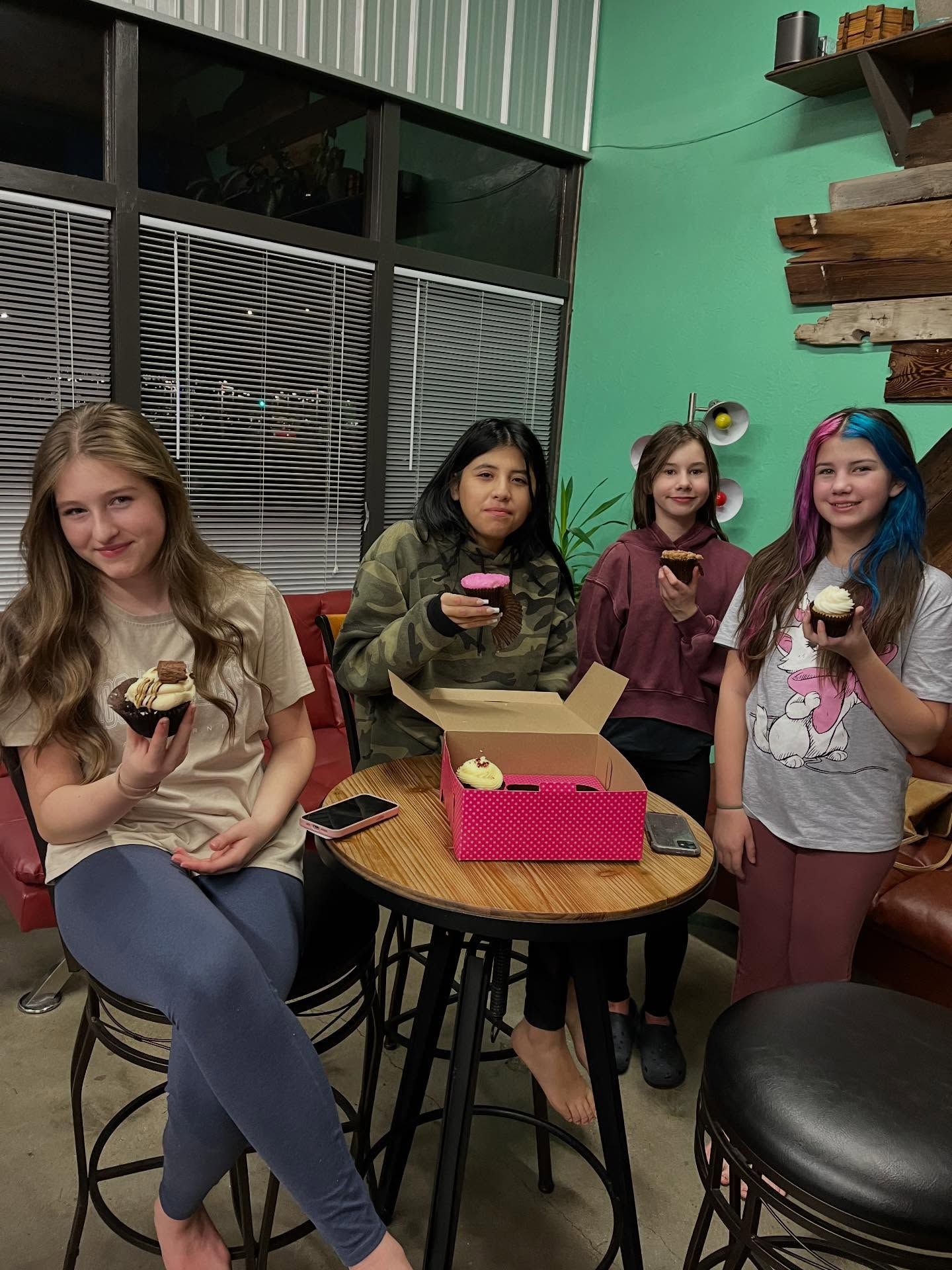 Four friends posing indoors with cupcakes, seated around a small table by a window.