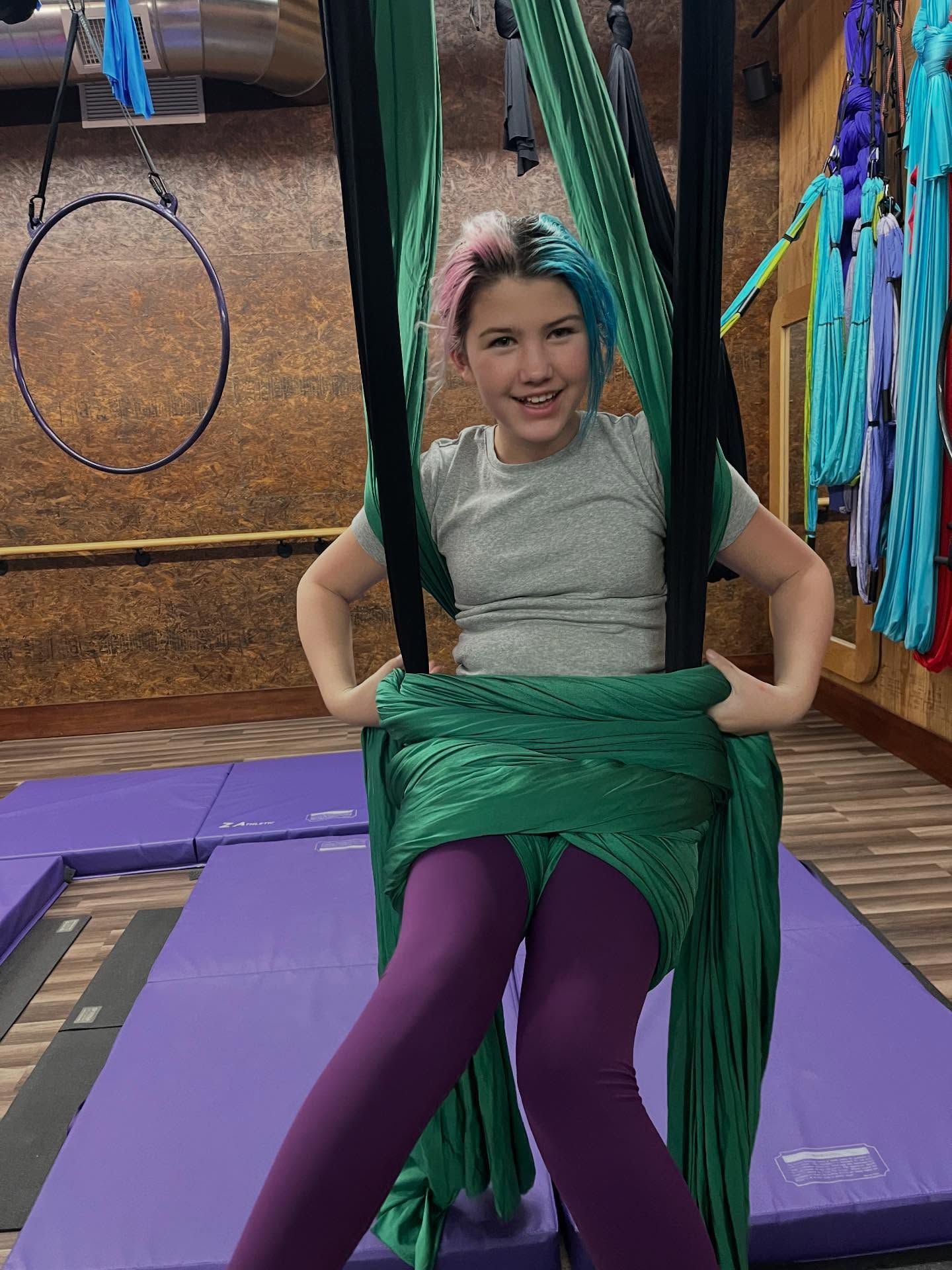 Person seated in a green aerial sling on a purple mat, smiling in an indoor gym with hanging hoops.