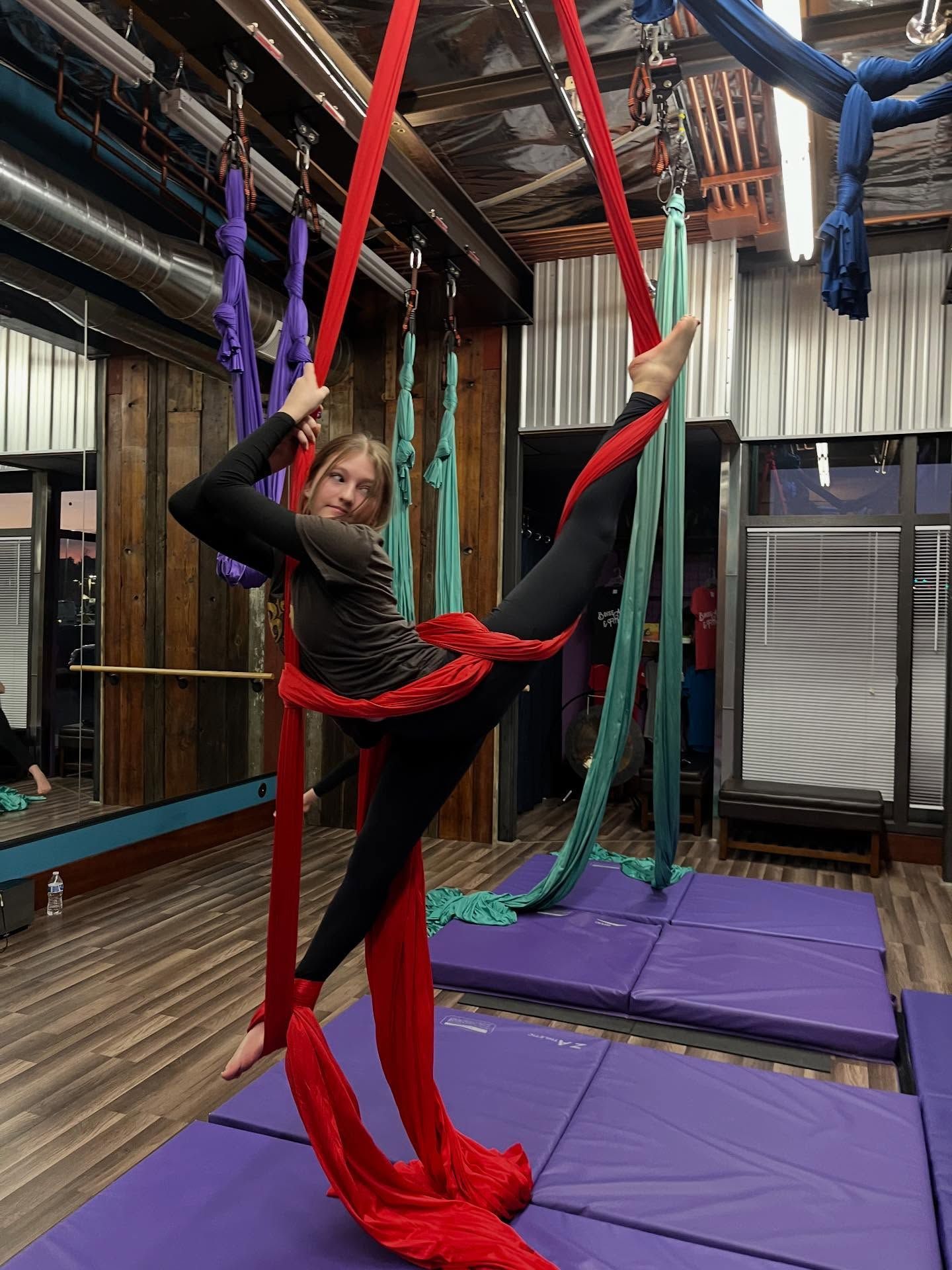 Person performing aerial silk pose in a gym, wrapped in red silks above purple mats.