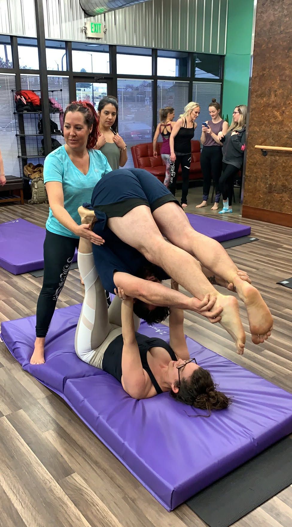 Two people practice acroyoga on purple mats while others watch in a studio
