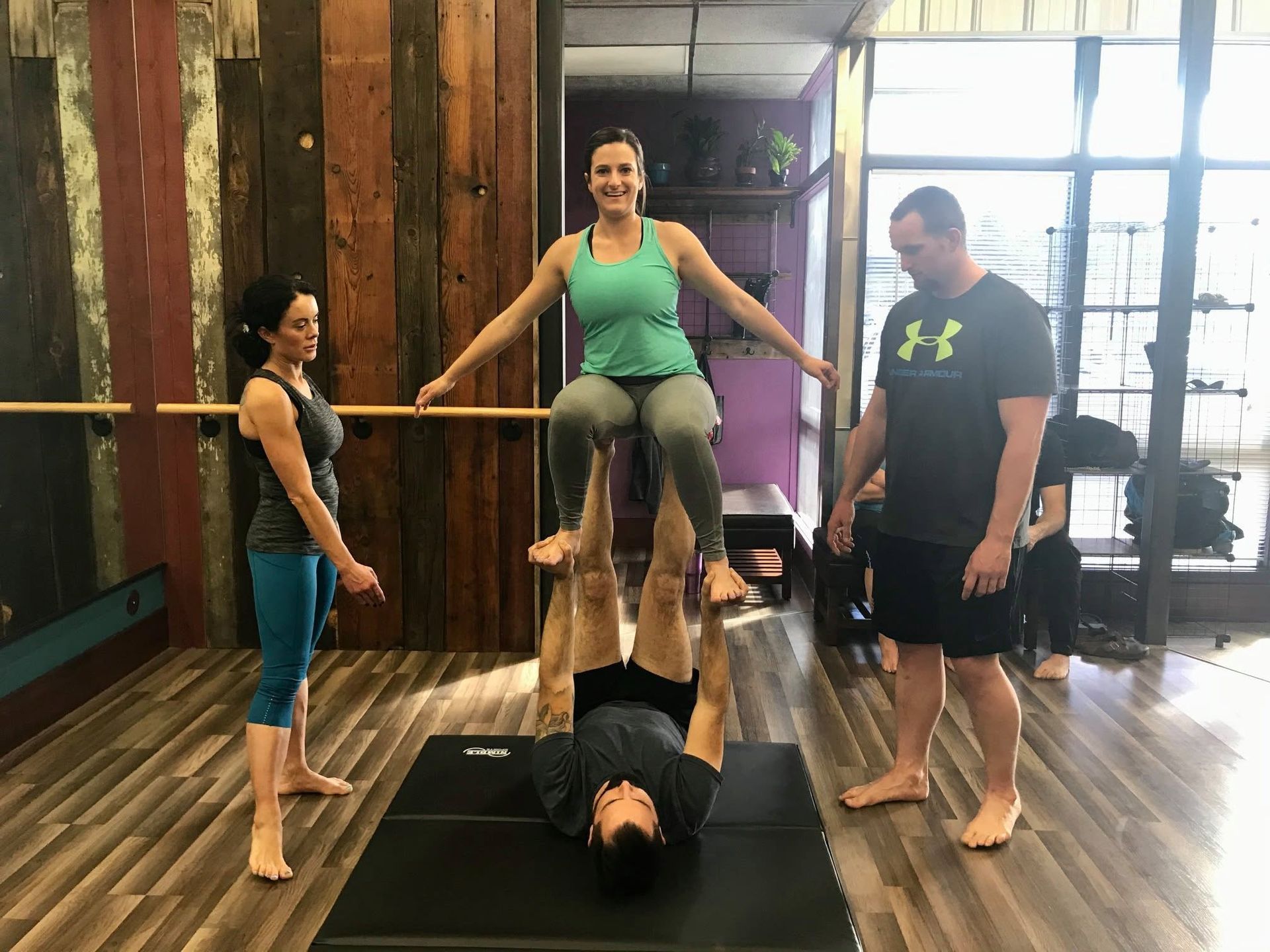 Four people doing an acro yoga pose in a gym studio with wooden floors and large windows