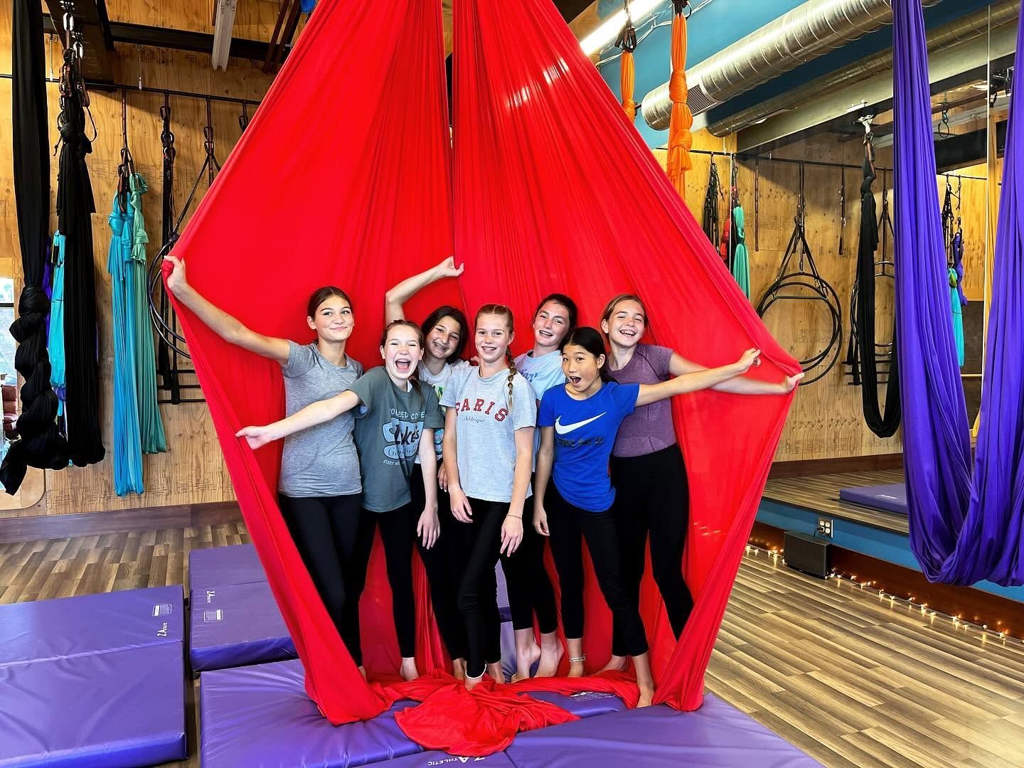 Group posing in a red aerial silk hammock at an indoor studio, with colorful hanging fabrics around them