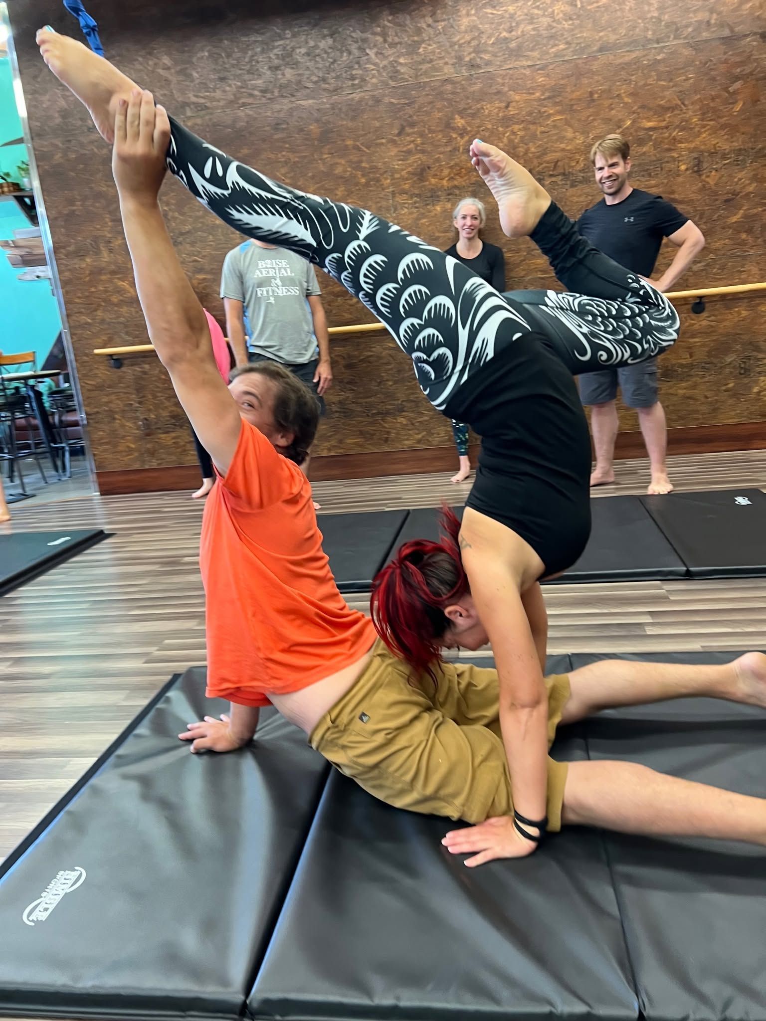 Two people perform a yoga acro pose on mats while two others watch in the background.