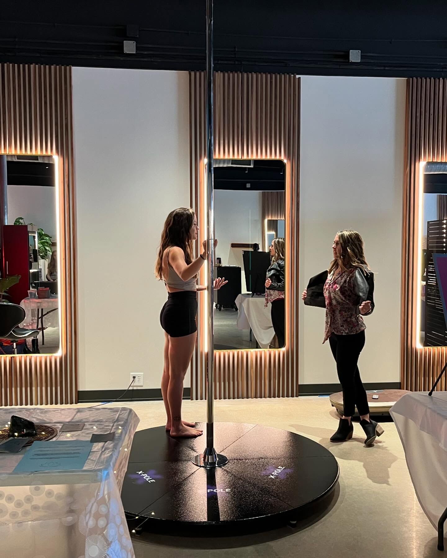 Two women stand on a mirrored pole-dance platform in a studio with tall mirrors and vertical wood slats.