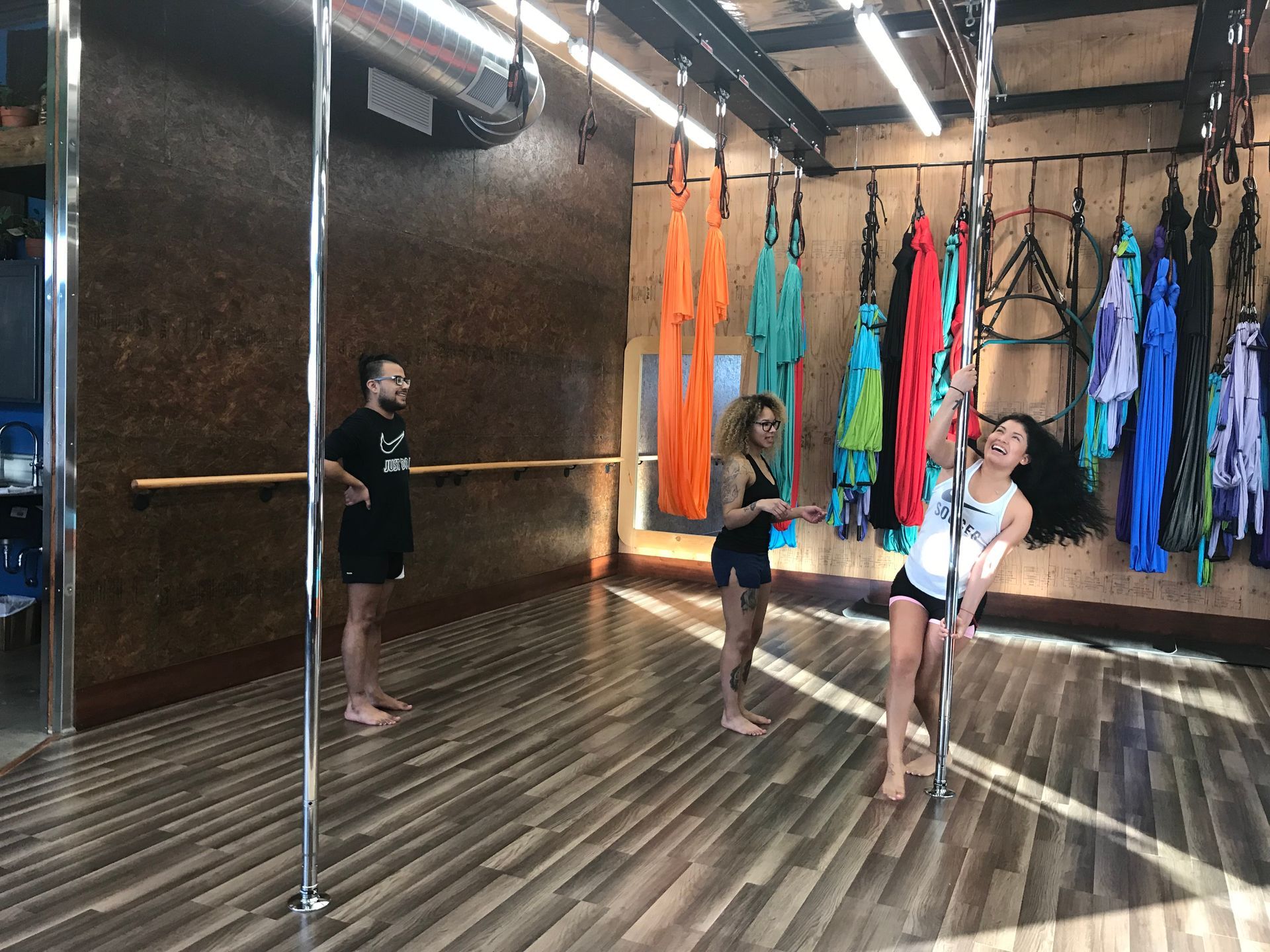 Three people stretching in a colorful aerial yoga studio with hanging silks and mirrors