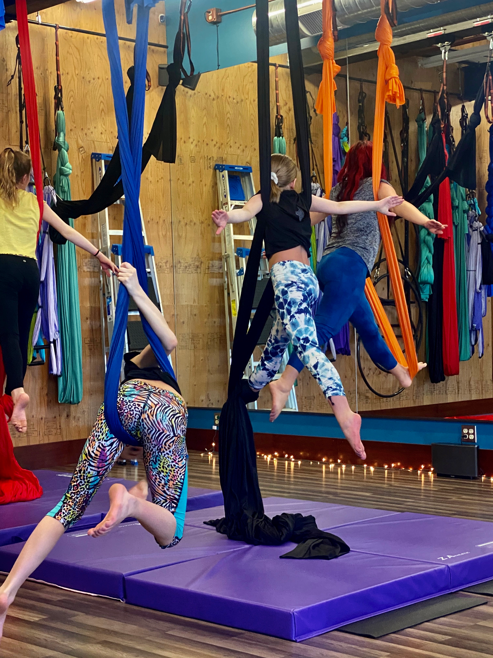 Three people suspended from colorful aerial silks in a studio, balancing on a purple mat.
