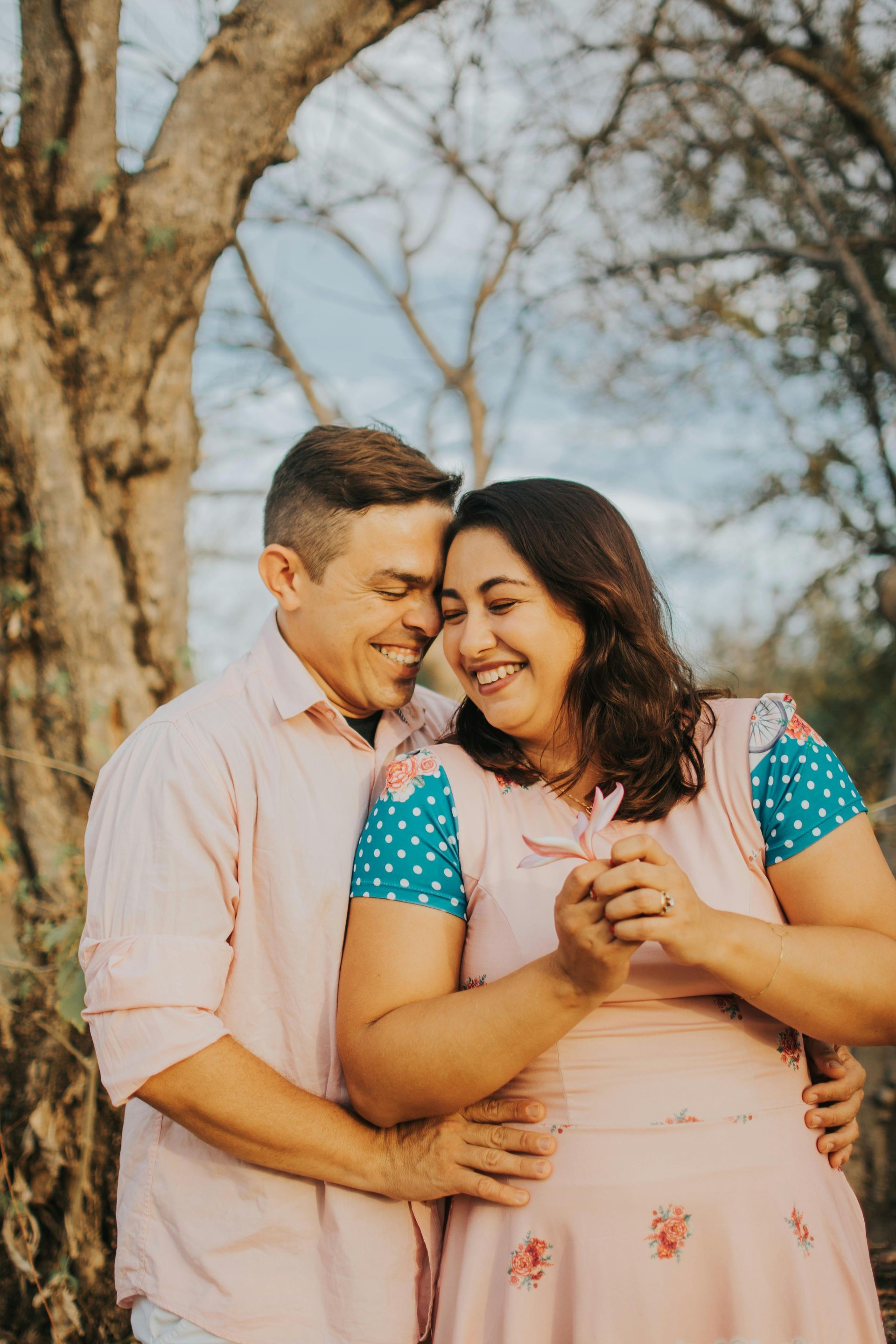Couple embraces, smiling. Man in pink shirt, woman in pink dress, outdoors with trees in the background. Level Up Falcon