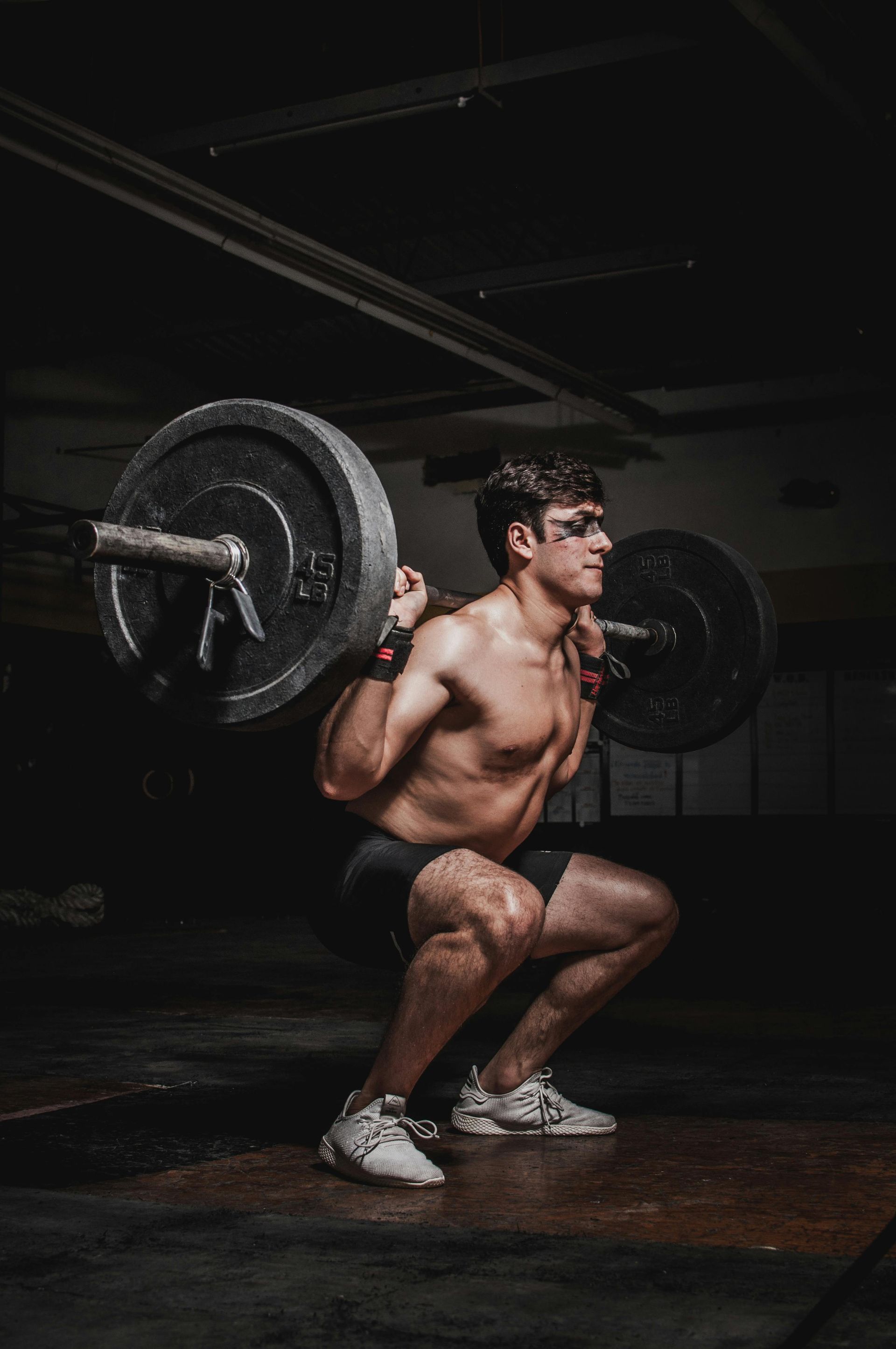 Man performs barbell squat in a dimly lit gym. He is shirtless with a determined expression. Level Up Falcon