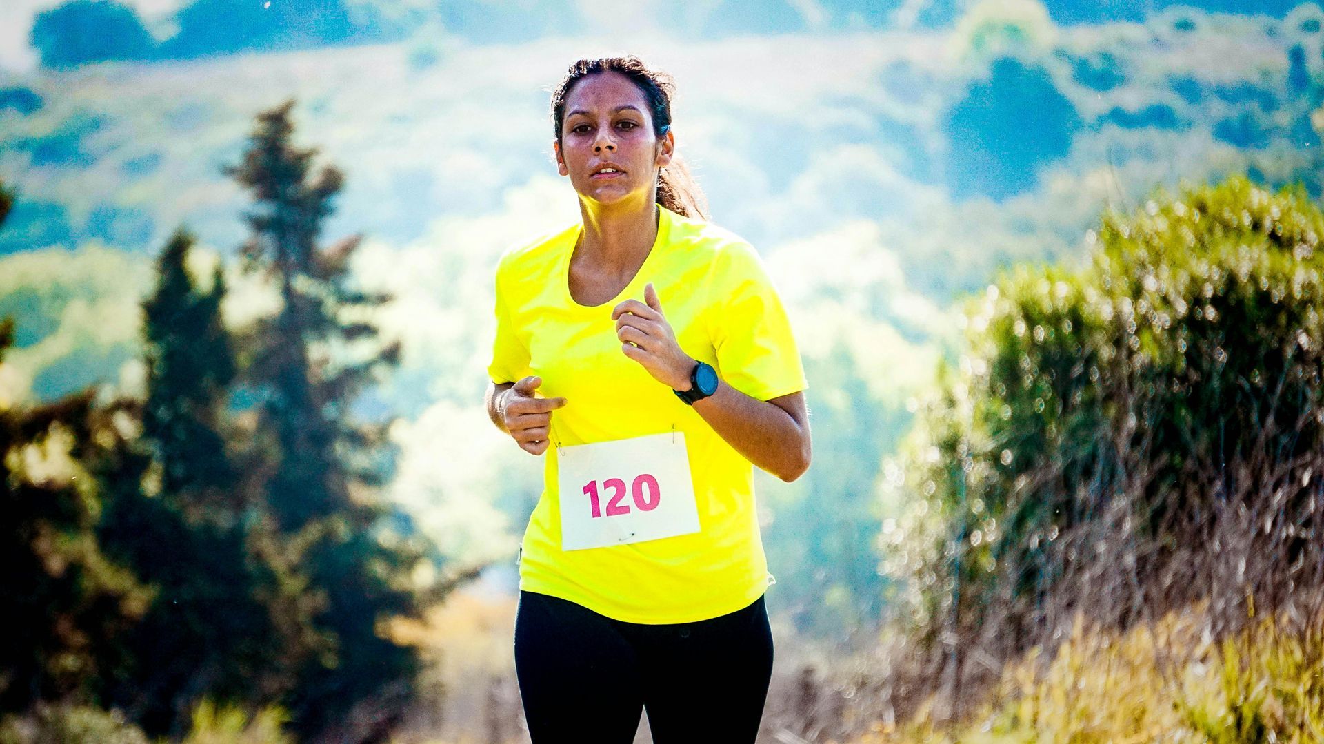 Woman running a race, wearing a yellow shirt and bib number 120. Set against a blurred, hilly landscape. Level Up Falcon