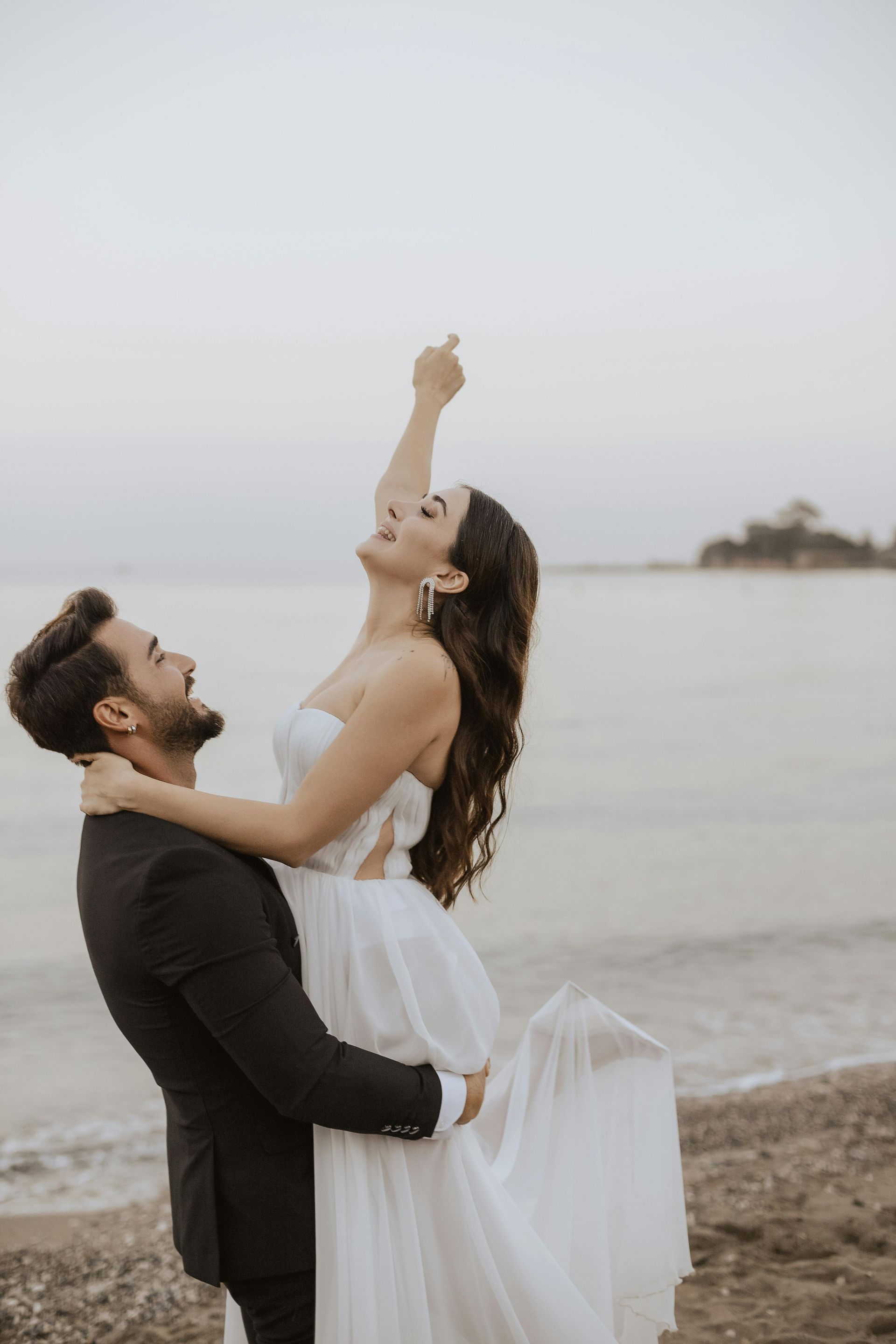 Man holding woman in a wedding dress on a beach, woman pointing upwards, both smiling, sea in background. Level Up Falcon