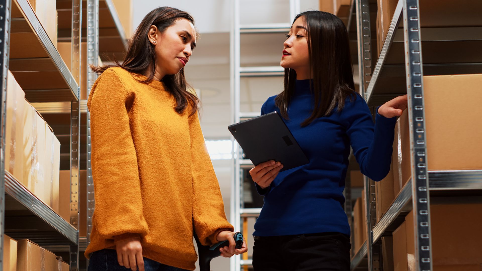Two women are standing in a warehouse looking at a clipboard.