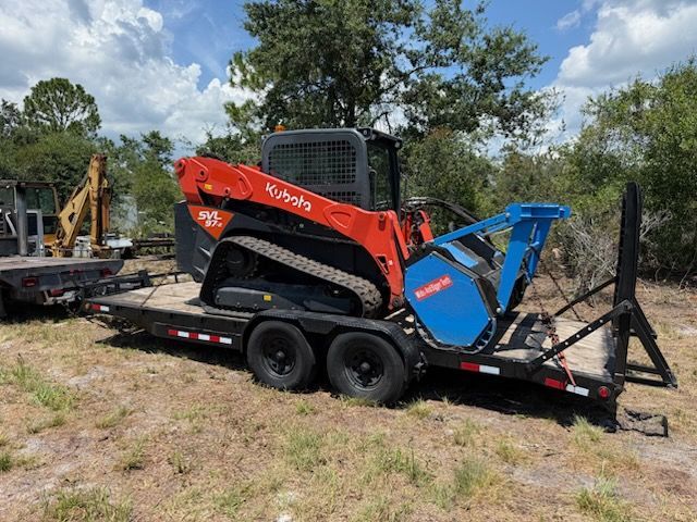 Orange-and-black Kubota skid steer on a trailer in a grassy field, with a blue attachment and trees behind